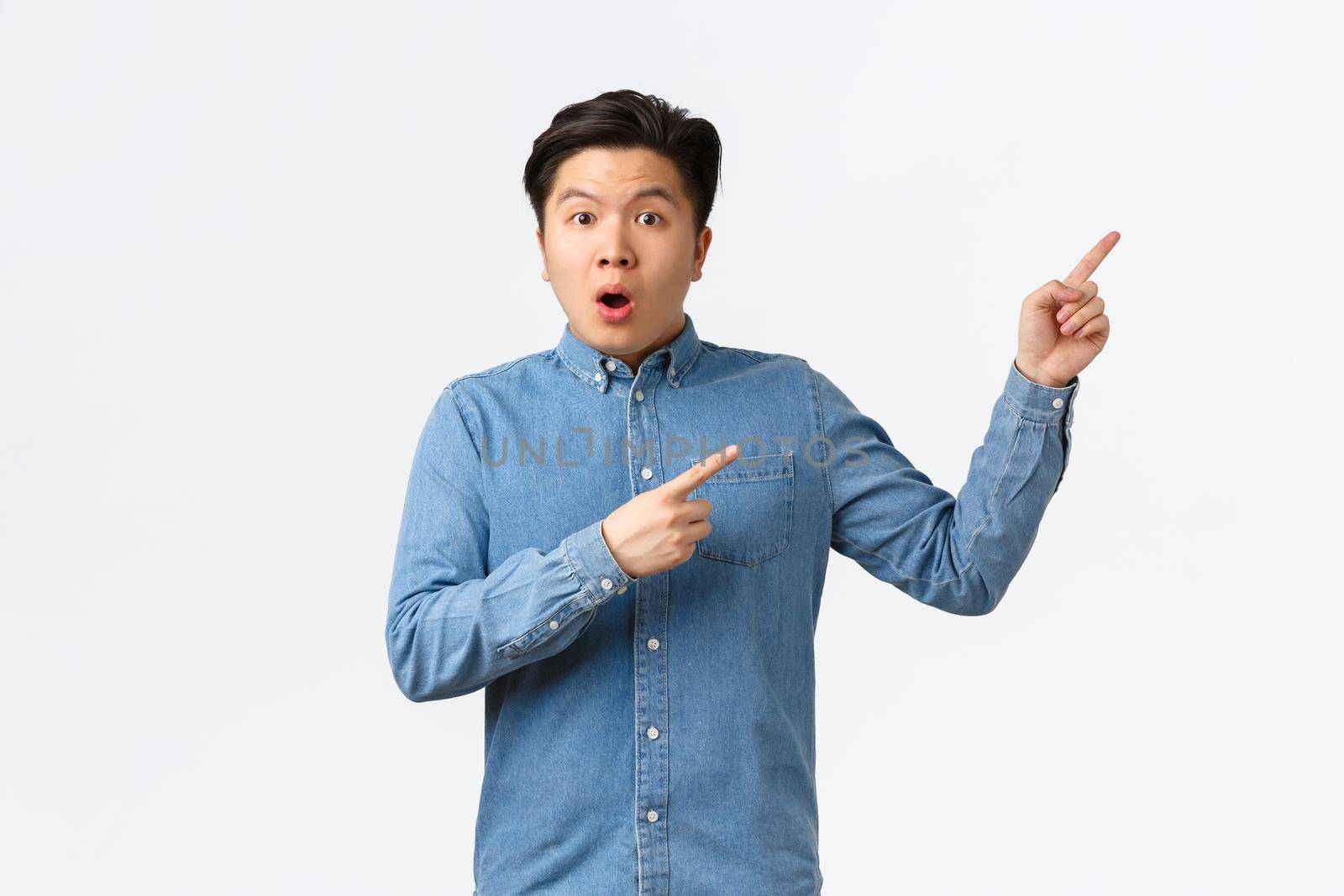 Shocked and astonished asian man asking question about product or announcement. Guy looking startled, pointing fingers upper right corner curious, discussing big event, white background by Benzoix