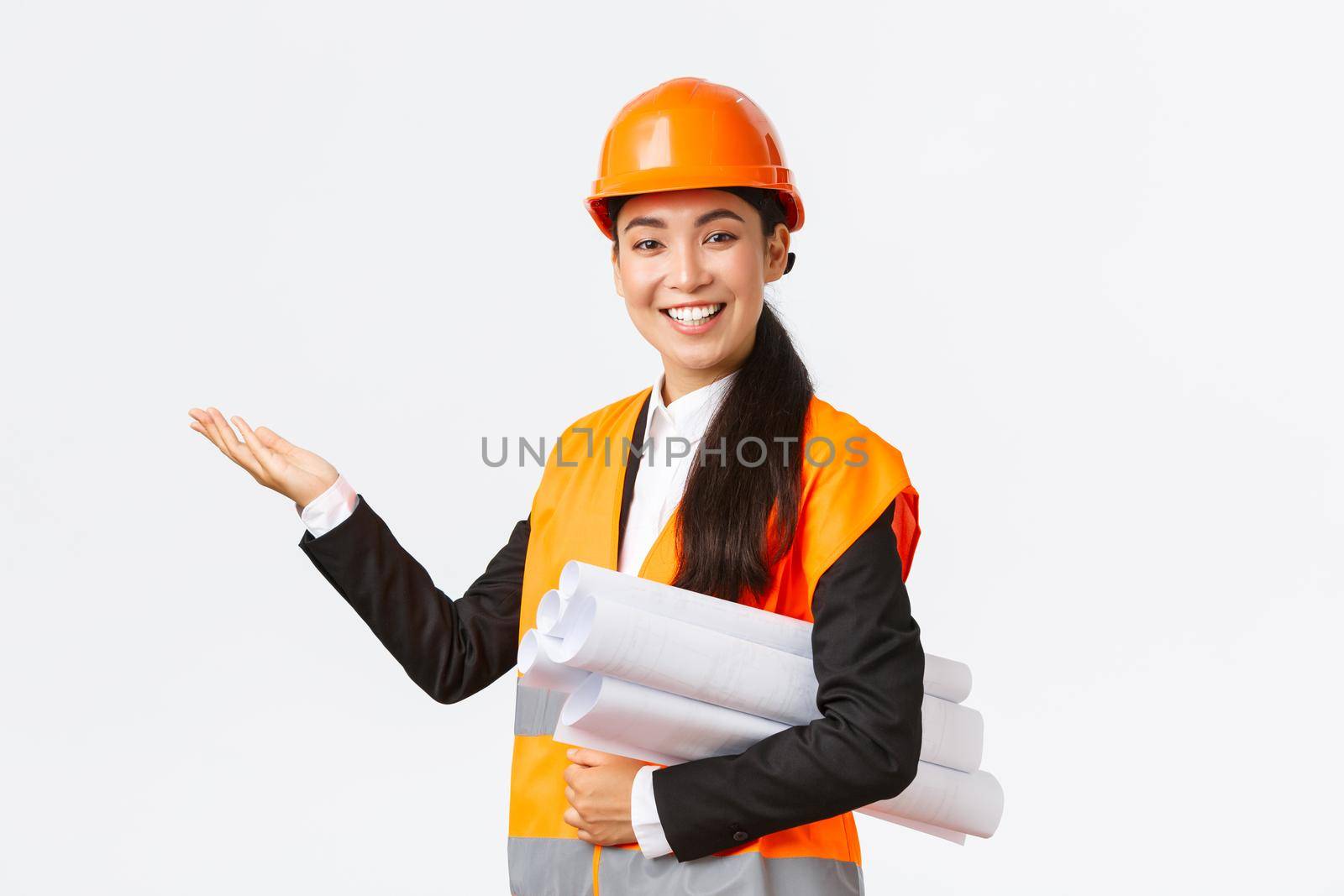 Smiling professional asian female architect, engineer in safety helmet introduce construction project, pointing hand left as carry blueprint, giving speech, introduce chart or plan of building.