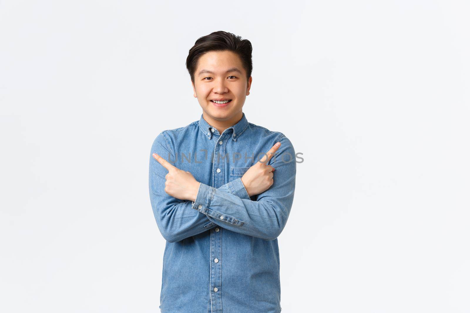 Smiling cheerful asian man with braces making announcement. Guy pointing fingers sideways at left and right variants, showing few options, recommend stores, standing white background by Benzoix