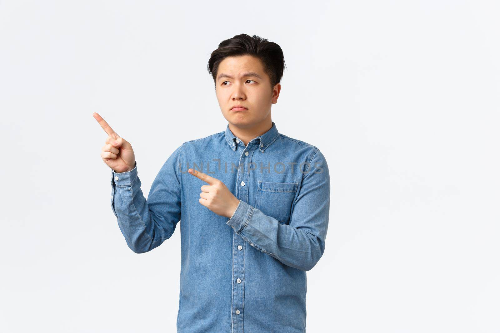 Unamused and disappointed asian gloomy man in blue shirt, frowning upset and pointing fingers upper left corner, complaining bad product, feeling distressed, standing white background.