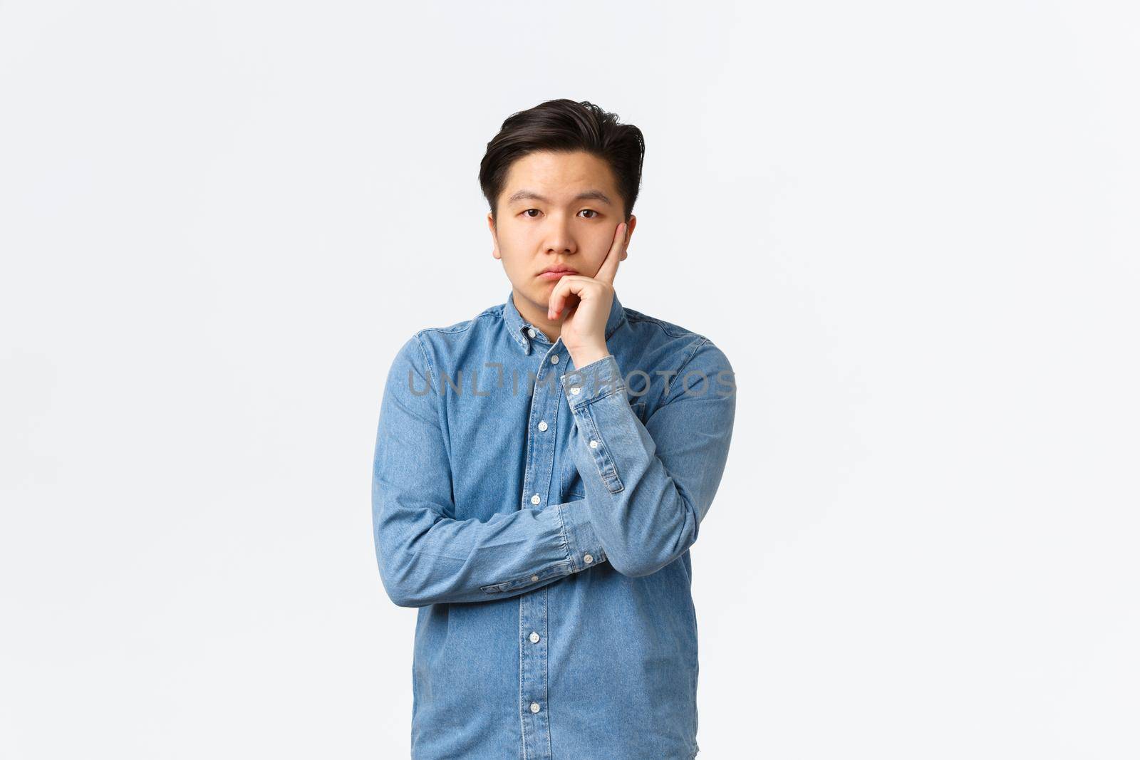 Bored and uninterested asian male in blue shirt, looking unamused and careless at camera, listening boring speech, standing displeased and annoyed over white background, tired of person.