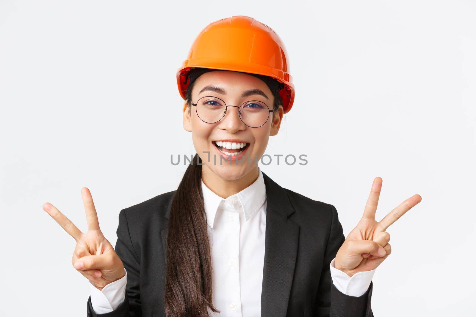 Close-up of cheerful successful female asian engineer, construction architect in safety helmet and business suit showing peace signs and smiling kawaii, standing white background.