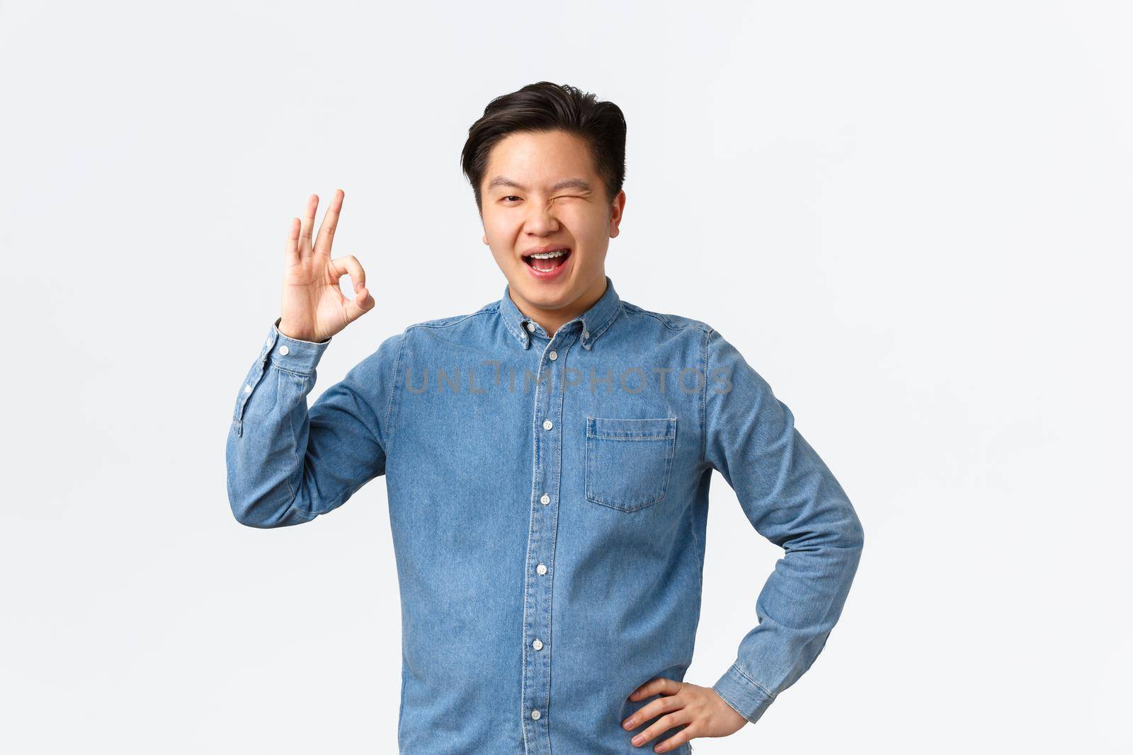 Cheerful confindent asian man have all under control, wink encouraging and show okay gesture, approve plan, congratulating person, saying well done, rate excellent job, standing white background by Benzoix