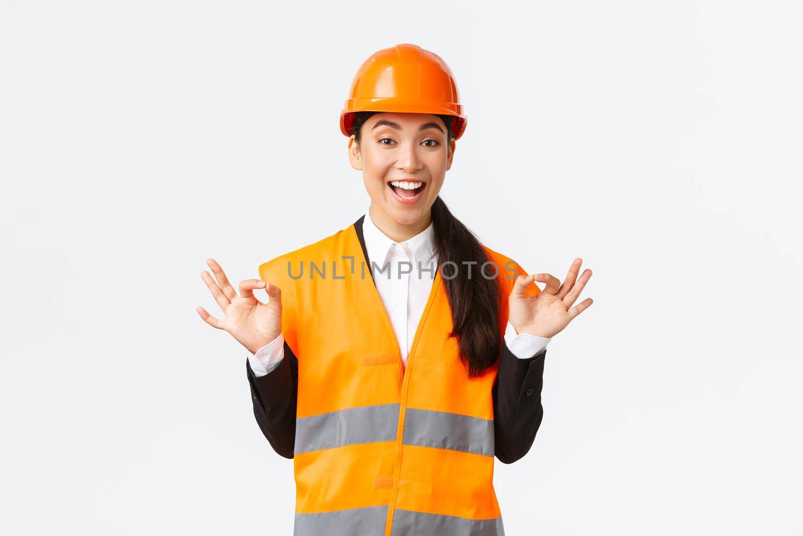 Excited and satisfied asian female chief engineer pleased with amazing work, showing okay gesture, assure clients, guarantee perfect quality, standing in safety helmet over white background.