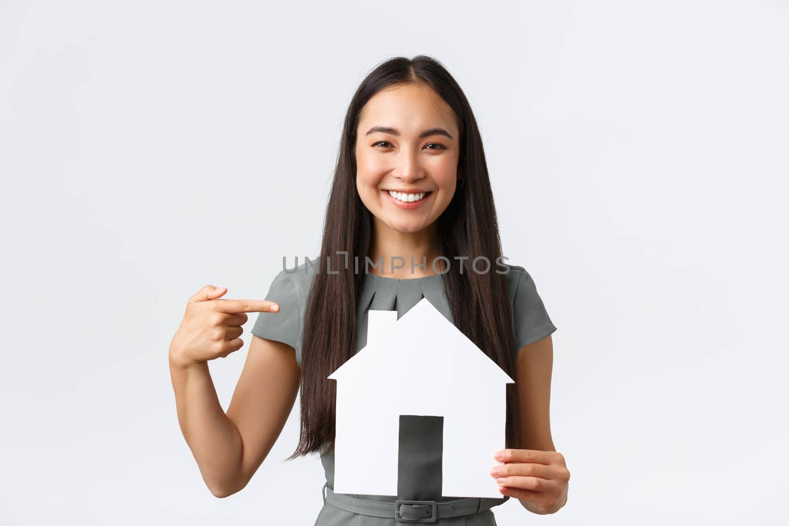 Insurance, loan, real estate and family concept. Smiling happy asian woman in dress, pointing finger at paper house in her hands, broker help find perfect apartment, standing white background.
