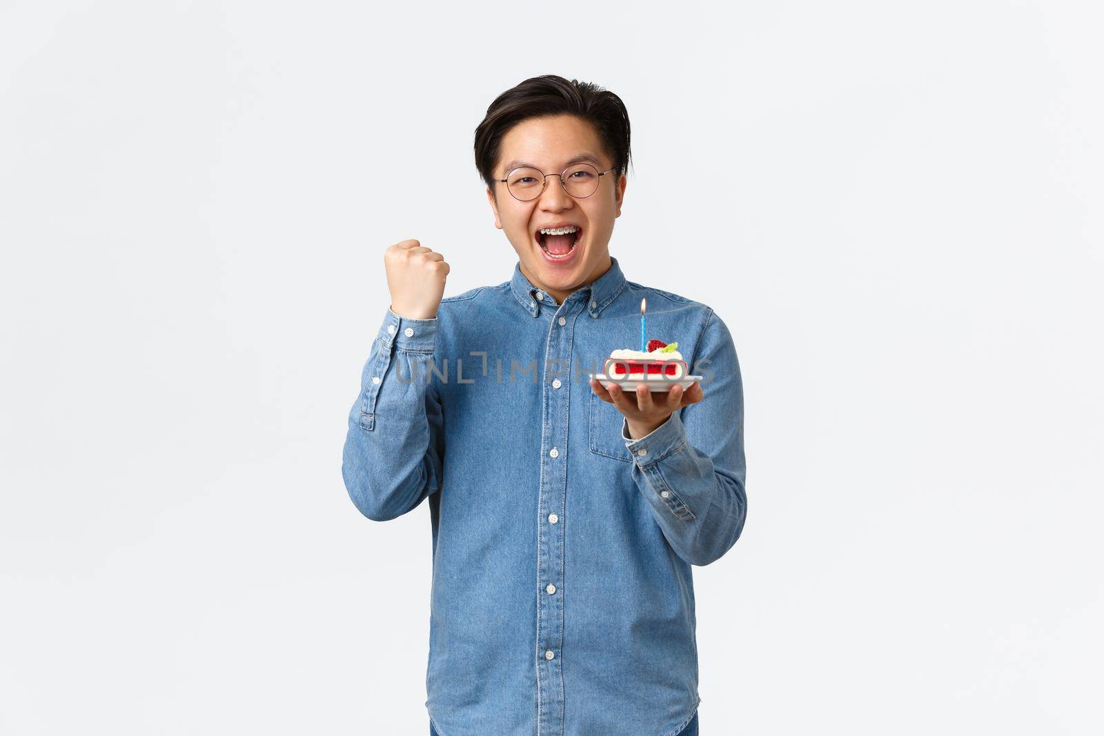 Celebration, holidays and lifestyle concept. Rejoicing happy asian man enjoying birthday party, holding b-day cake with lit candle and fist pump, triumphing and saying yes, assertive wish come true by Benzoix