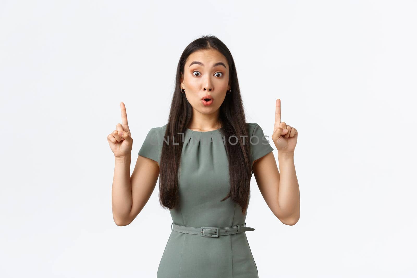 Small business owners, women entrepreneurs concept. Excited asian female in dress pointing fingers up and looking shocked at camera, making big announcement, white background.