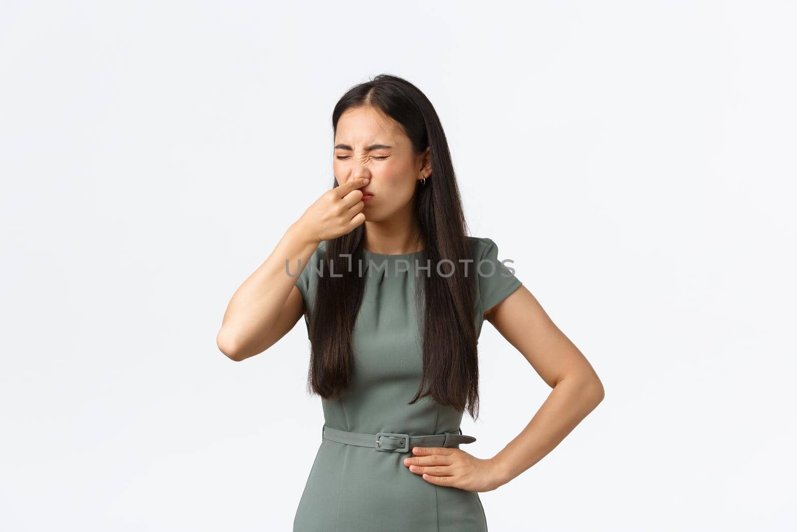 Small business owners, women entrepreneurs concept. Disgusted asian female in elegant dress shut her nose with fingers and grimacing from nasty reek, bad smell, standing white background.