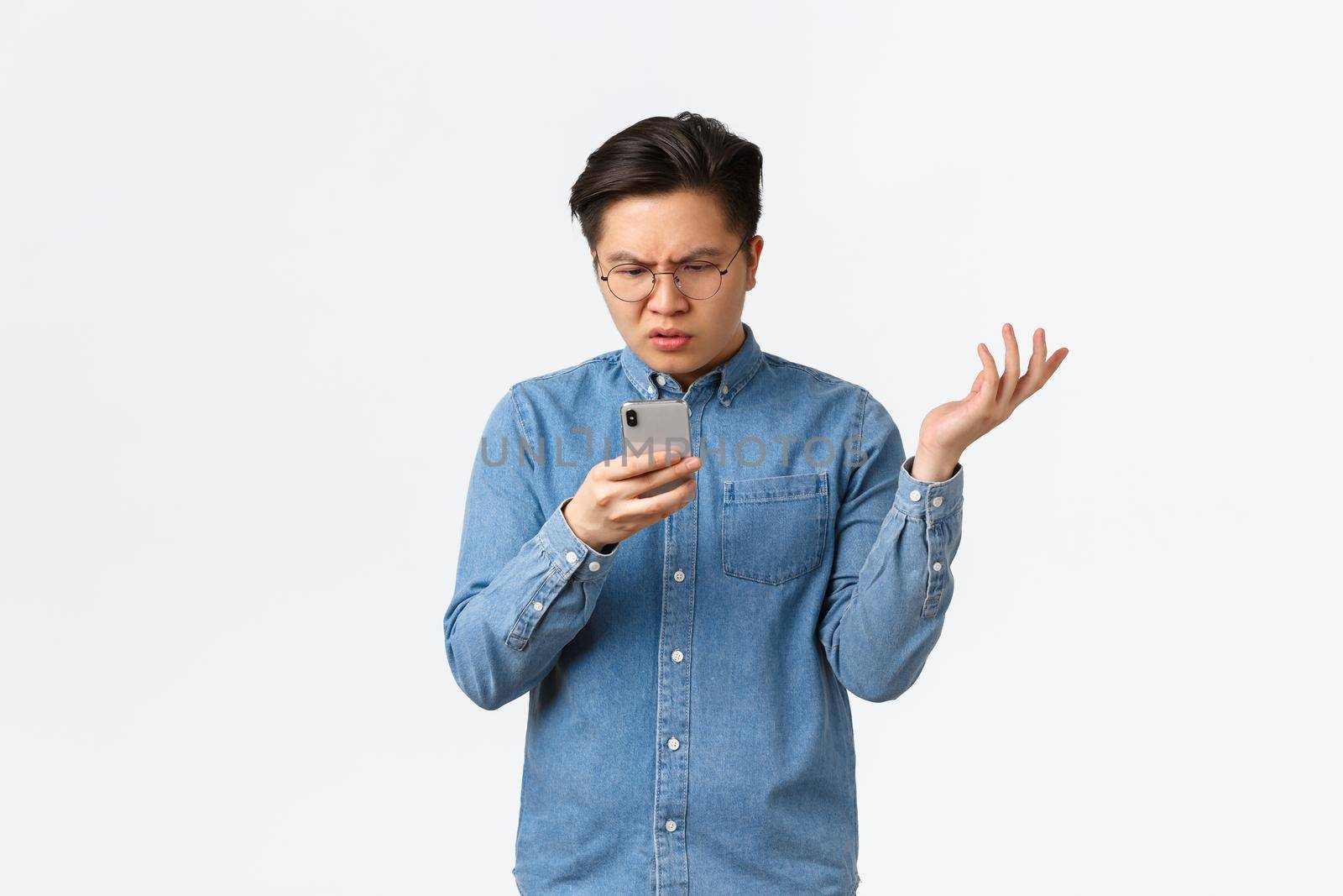 Frustrated and confused asian guy in glasses looking puzzled at mobile phone screen, raising hand up questioned, cant understand what happening online, standing white background perplexed.