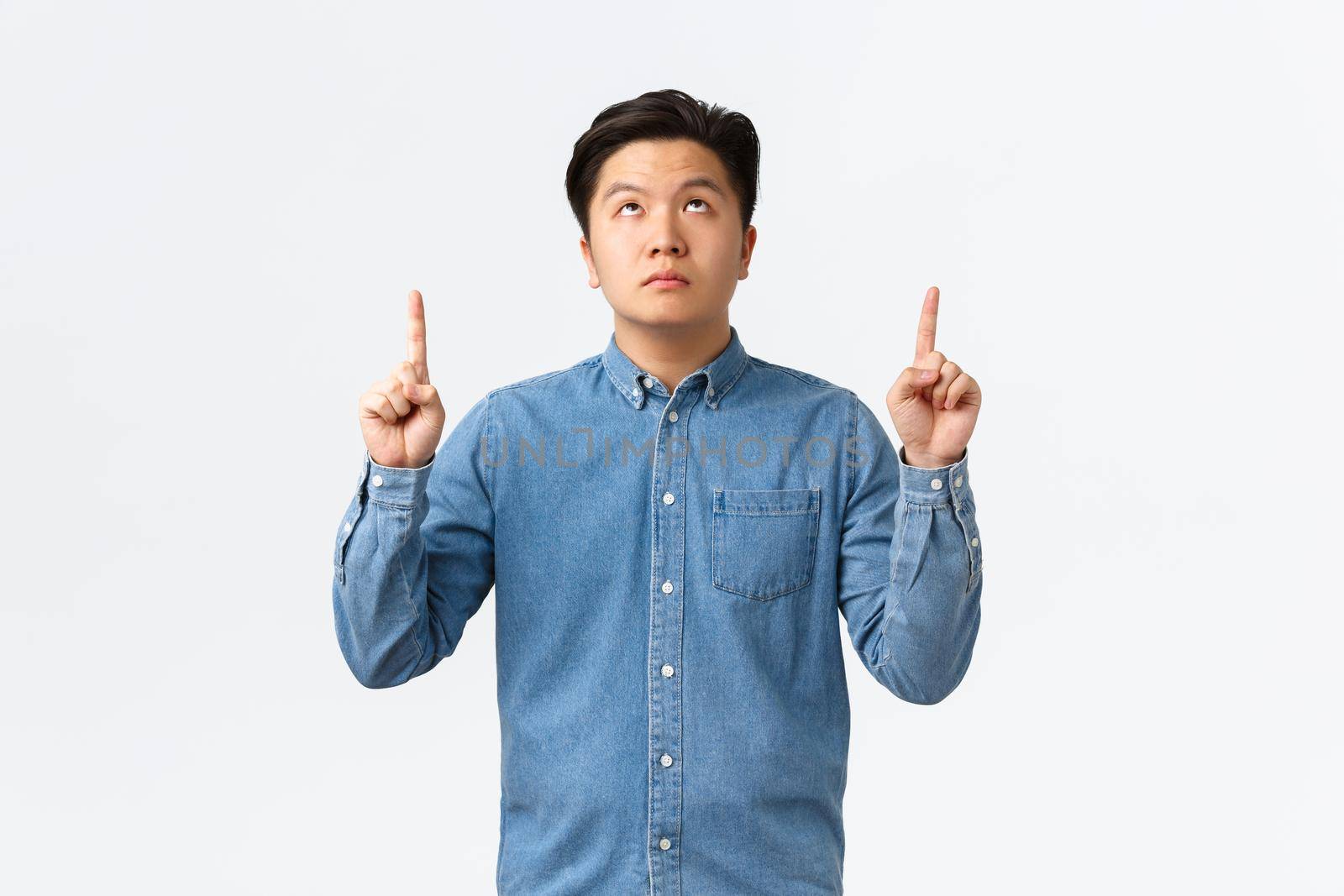 Unamused bored asian guy in blue shirt, looking and pointing fingers up with poker face, no emotions, being careless about information upwards, reading sign without interest, white background.