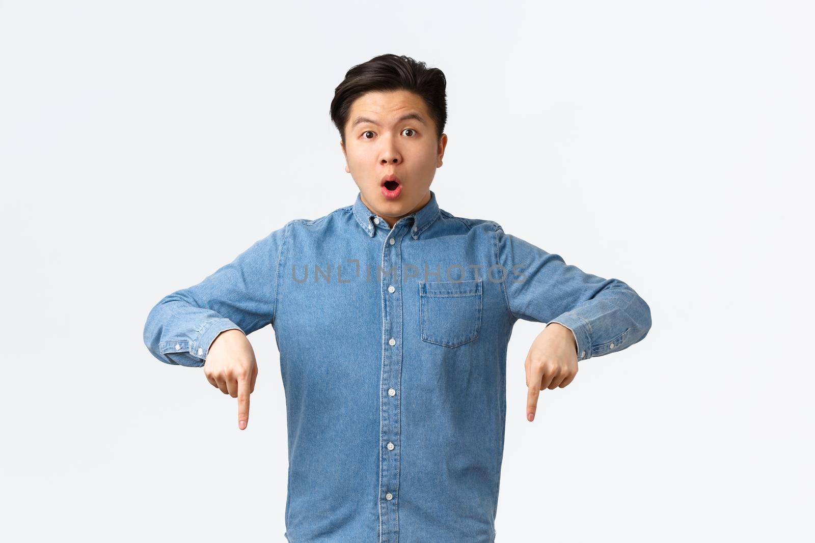 Shocked and impressed asian guy in blue shirt, pointing fingers down and looking at camera speechless, asking question about product, found something interesting, standing white background.
