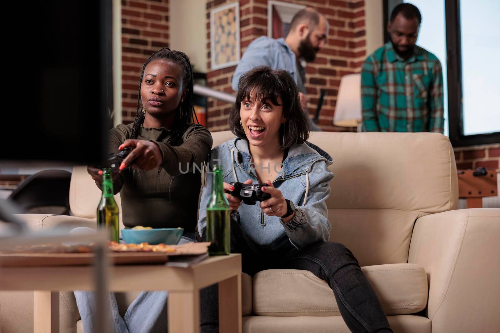 Multiethnic women playing video games with controller on console, having fun together at party gathering with friends. People enjoying leisure activity and competition, drinking beer.