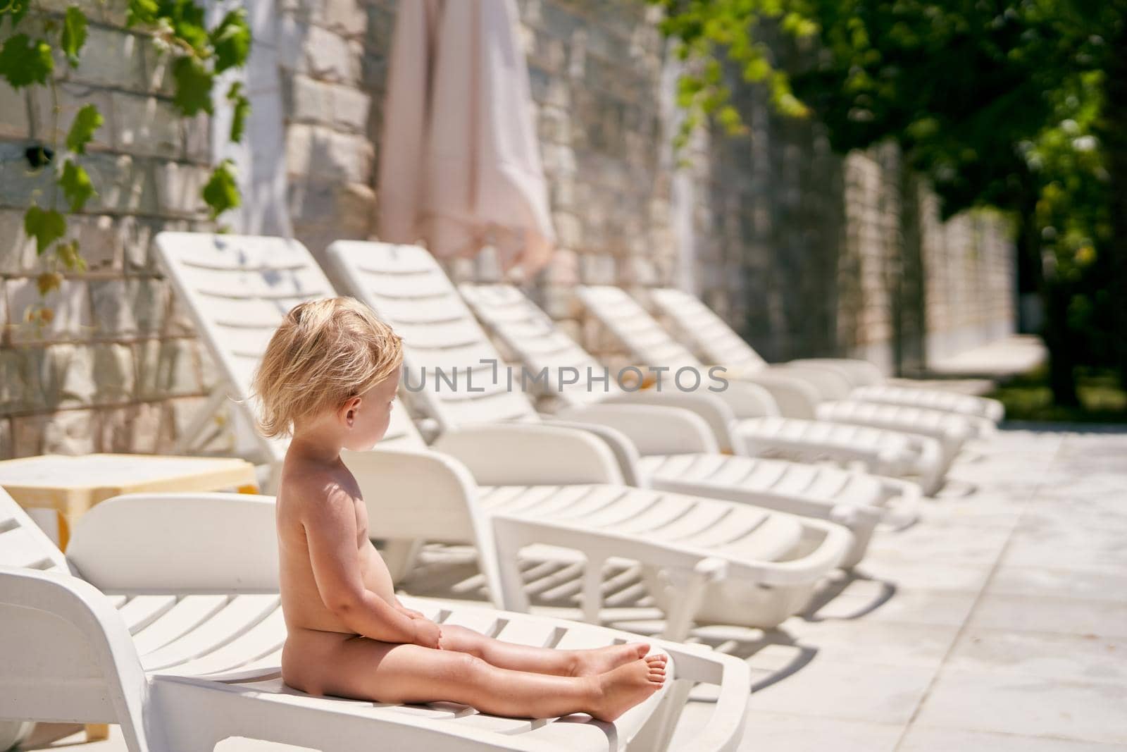 Little girl sits on a sun lounger near a stone wall by Nadtochiy