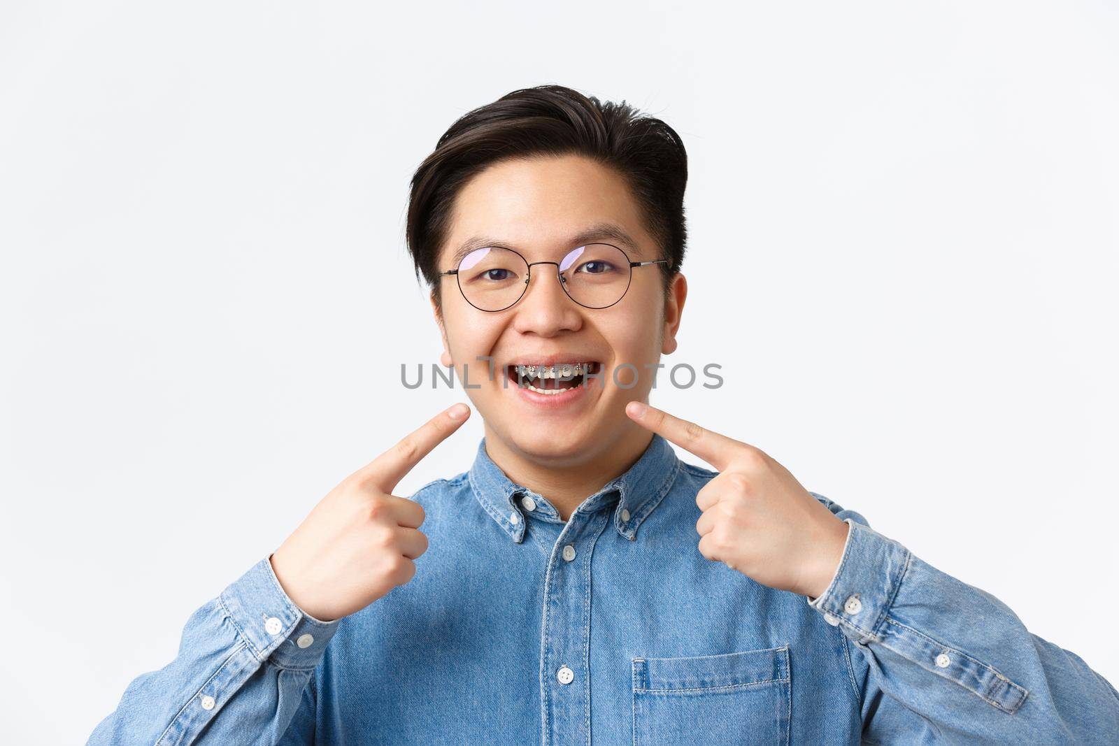 Orthodontics and stomatology concept. Close-up of satisfied asian guy, dental clinic client smiling happy and pointing at his dental braces, standing white background, recommend quality by Benzoix