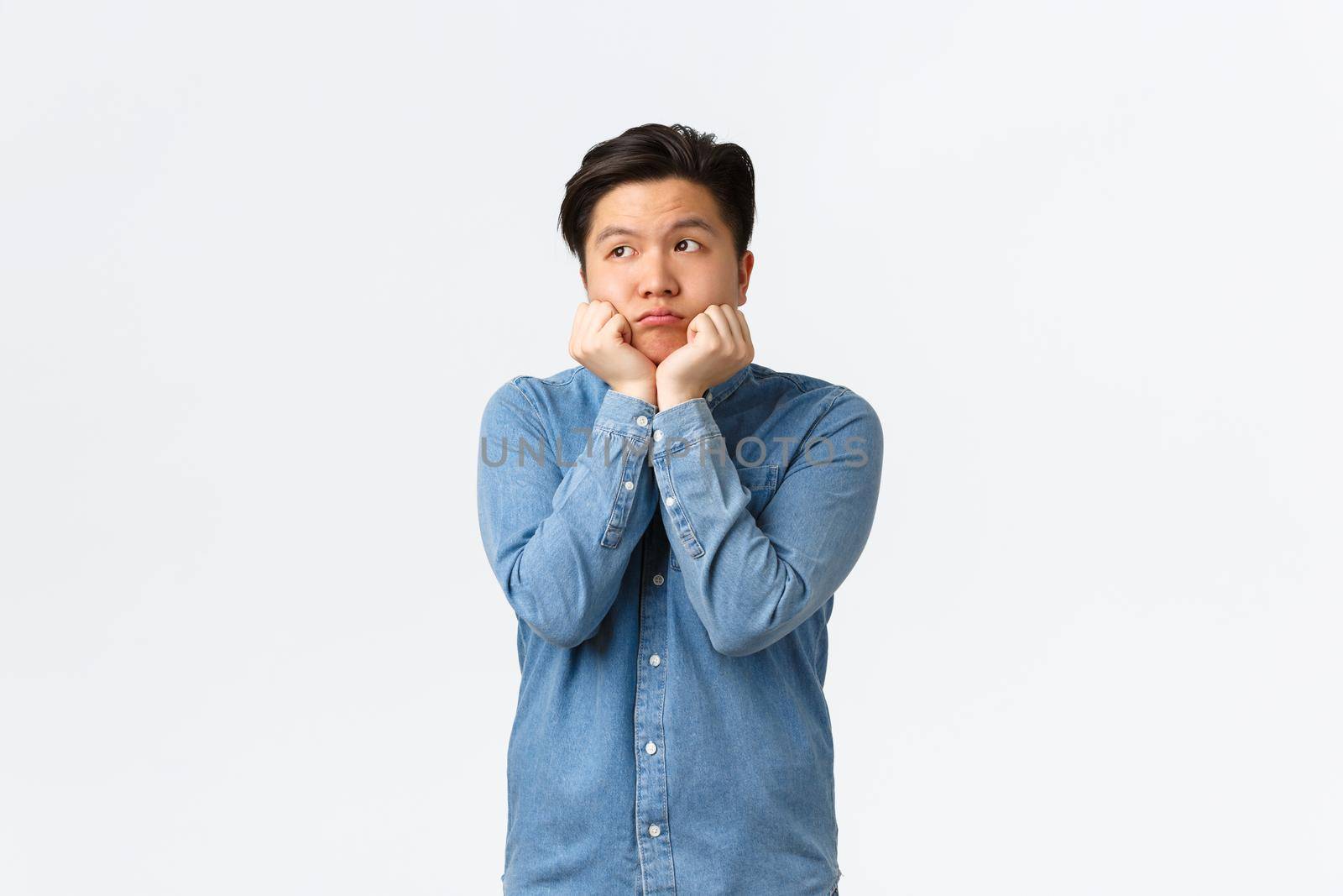Silly and cute dreamy asian guy looking upper left corner with longing face, leaning on palms timid, thinking about something he desires, feeling bored or sad, standing white background by Benzoix