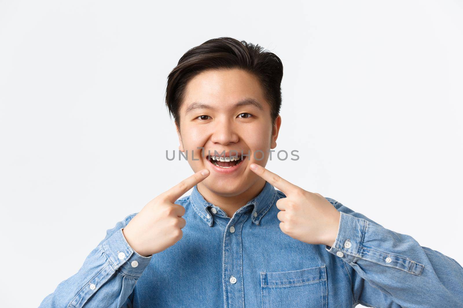 Orthodontics and stomatology concept. Close-up of happy smiling asian man pointing fingers at dental braces on teeth with pleased expression, recommend dentist clinic, standing white background by Benzoix