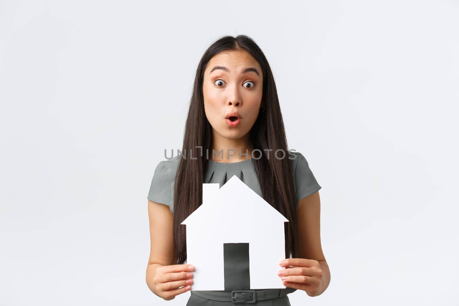 Insurance, loan, real estate and family concept. Surprised asian woman in dress, holding paper house in hands react to big announcement, found excellent apartment for cheap price, white background.