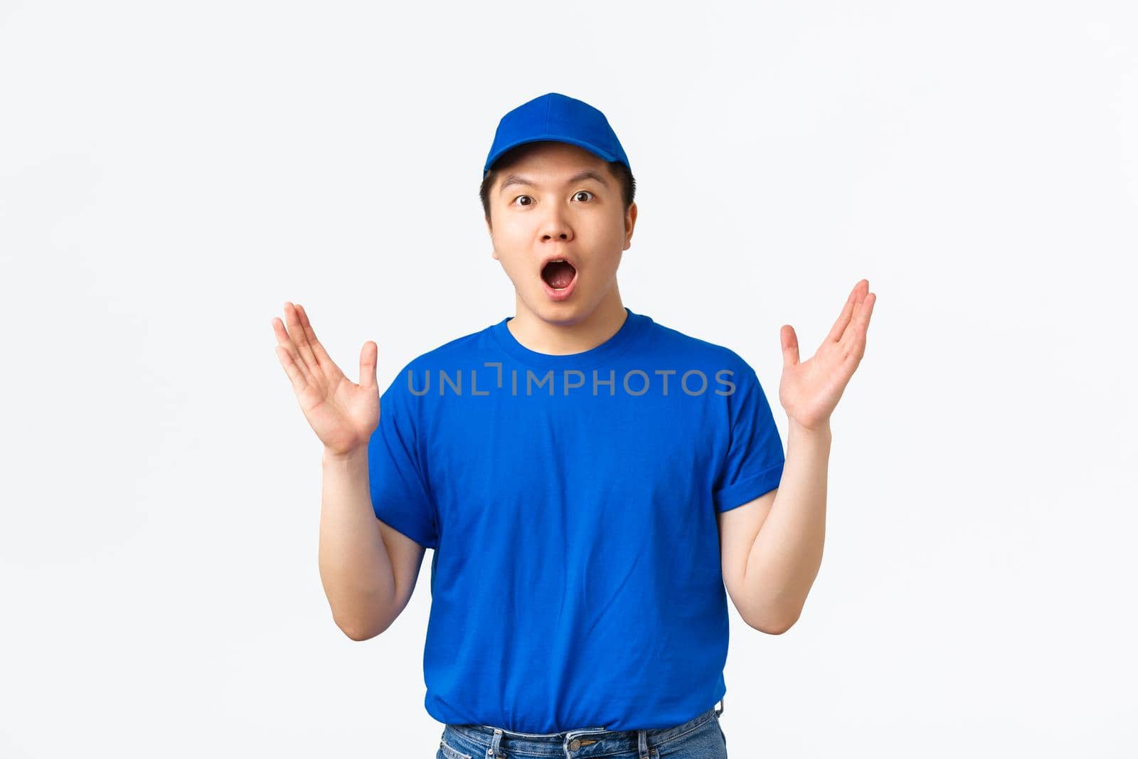 Surprised and excited courier in blue uniform react to awesome news, big announcement in mail company. Impressed delivery guy shaking raised hands and gasping amazed, white background.