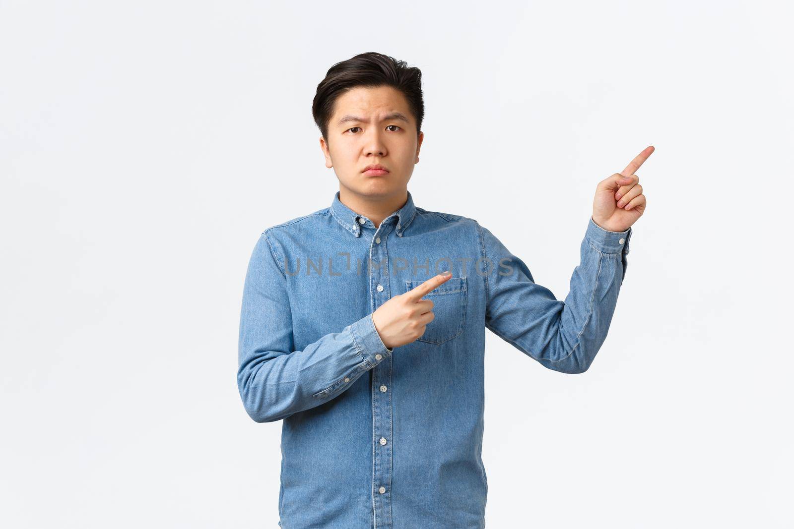 Suspicious and doubtful asian male in blue shirt, frowning skeptical and displeased, pointing fingers upper right corner, asking question, complaining on something bothering, white background by Benzoix