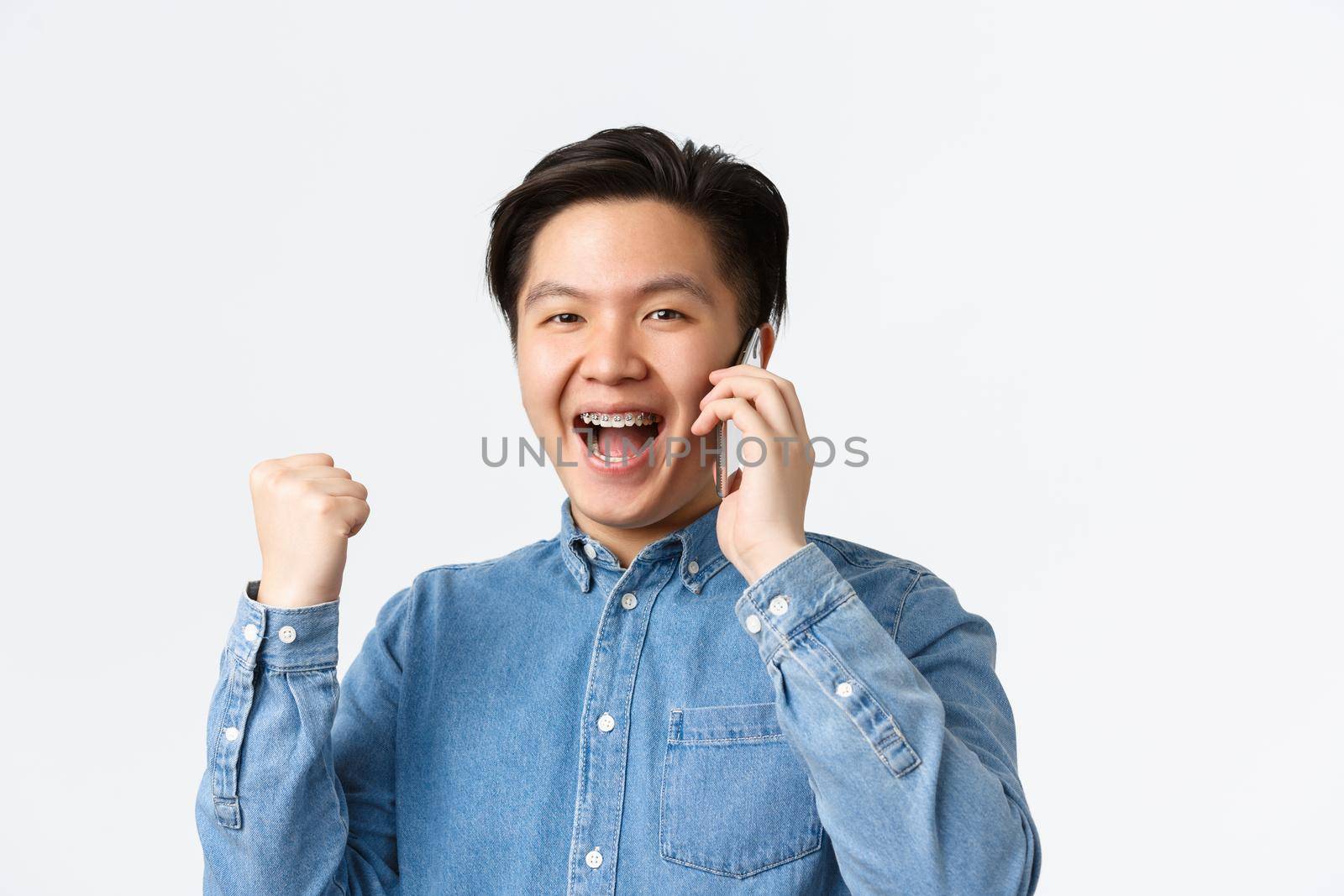 Close-up of happy handsome asian guy rejoicing great news, talking on mobile phone and fist pump, triumphing and saying yes, achieve goal. Man receive call from employer, got job, white background.