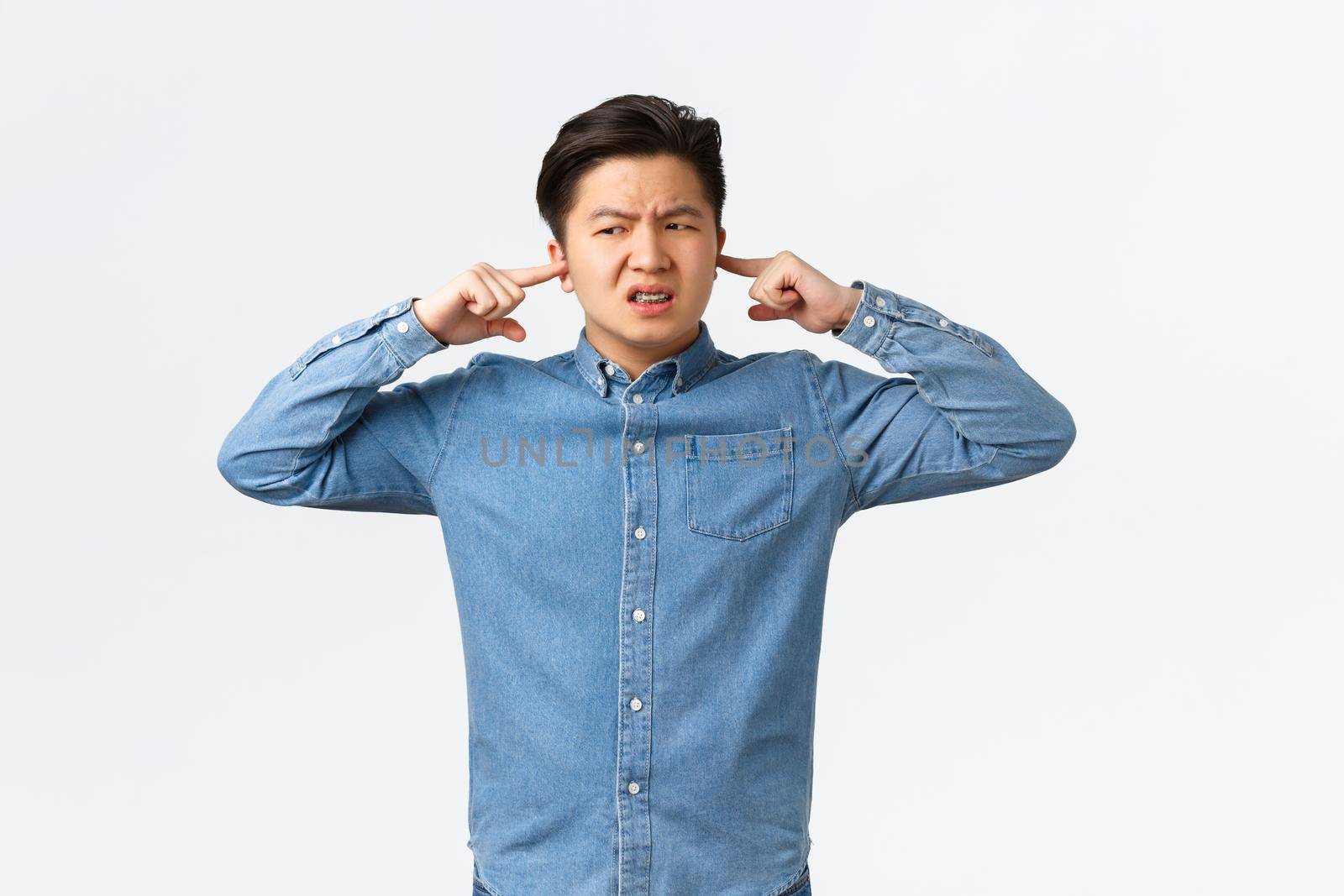 Disturbed and frustrated asian man complaining loud noise, shut ears and looking left displeased, frowning upset, telling neighbour be quiet, college student cant focus because of noisy awful music.