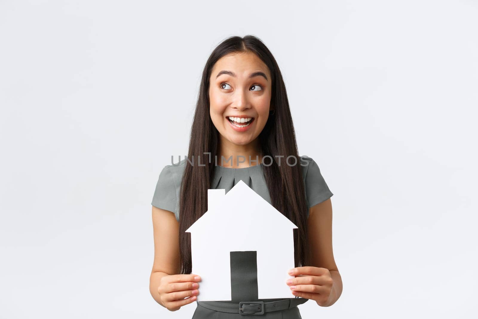 Insurance, loan, real estate and family concept. Smiling happy asian woman found perfect home, holding paper house and rejoicing, looking amused upper left corner at banner, white background.