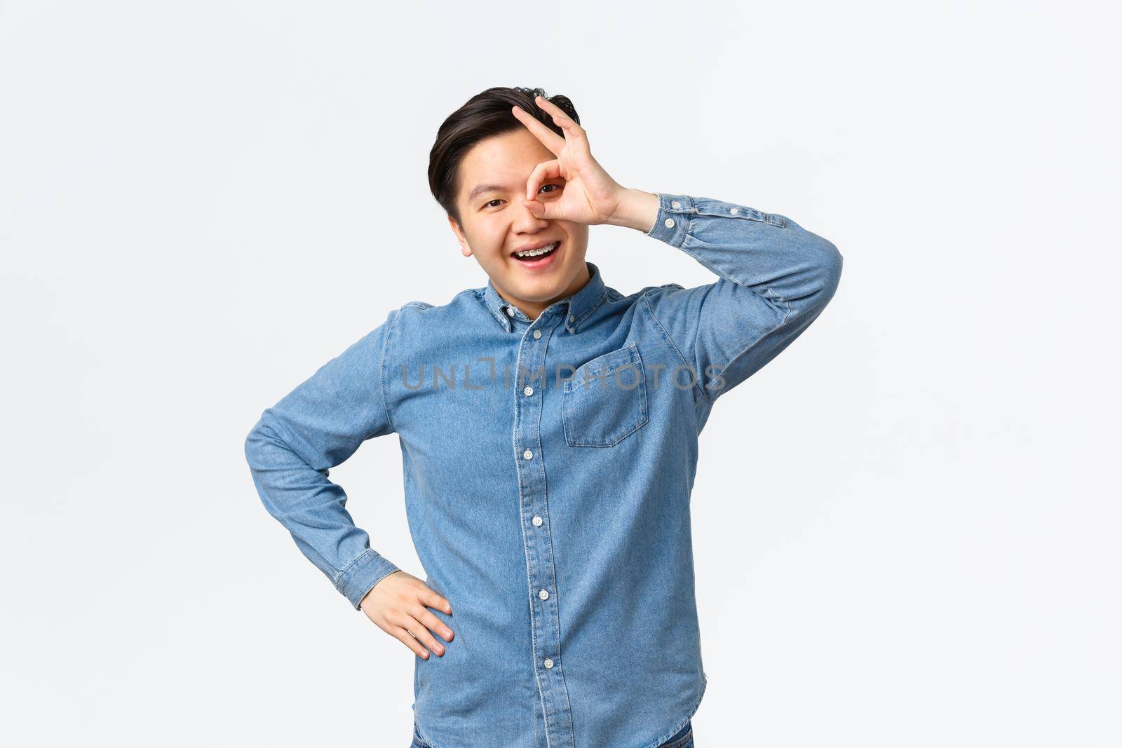 Smiling cheerful and satisfied asian man showing okay gesture over eye, looking through with pleased expression, assure everything good, recommend excellent service, guarantee.