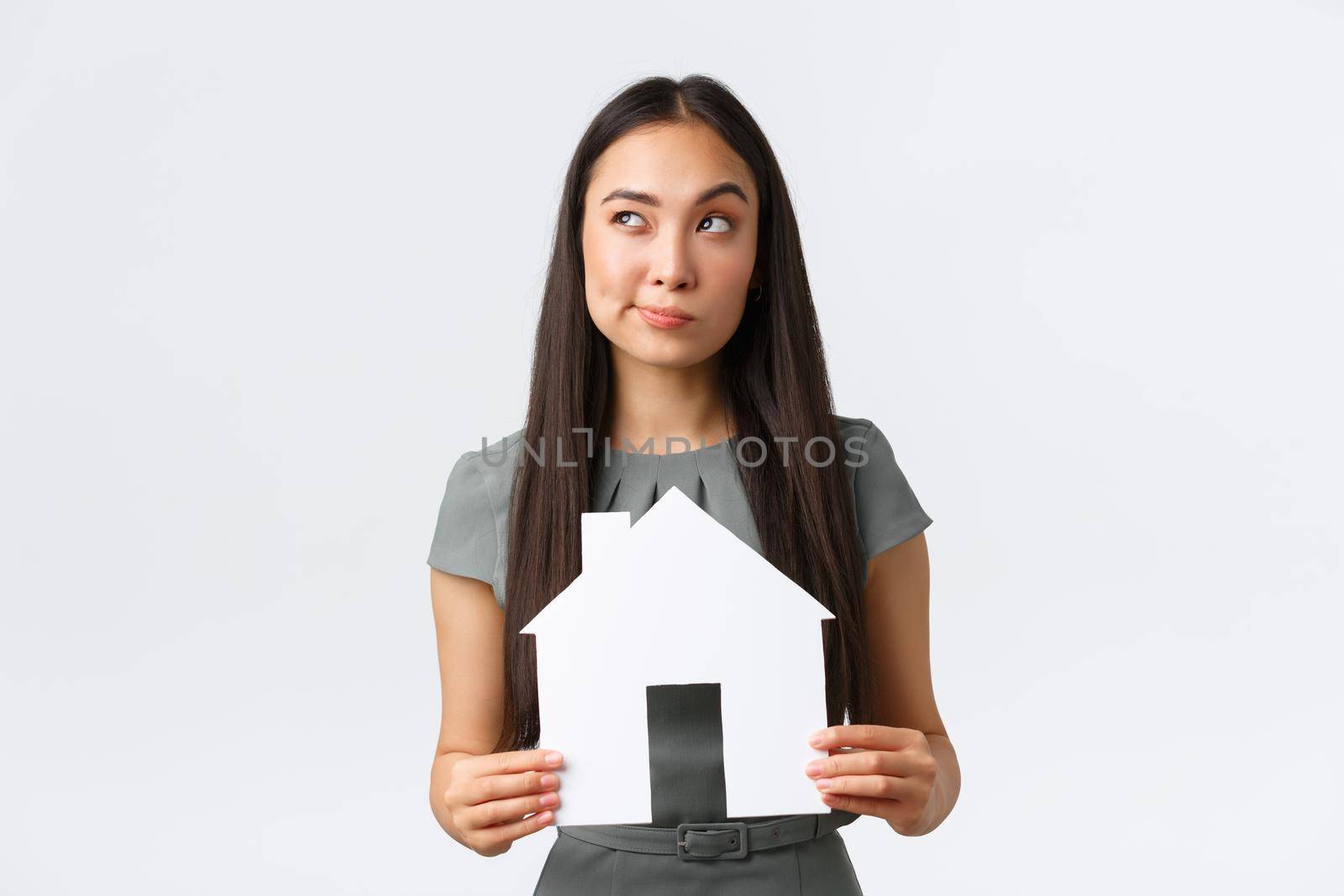 Insurance, loan, real estate and family concept. Thoughtful and doubtful asian woman thinking about buying new apartment, holding paper house and looking away, pondering, making decision.
