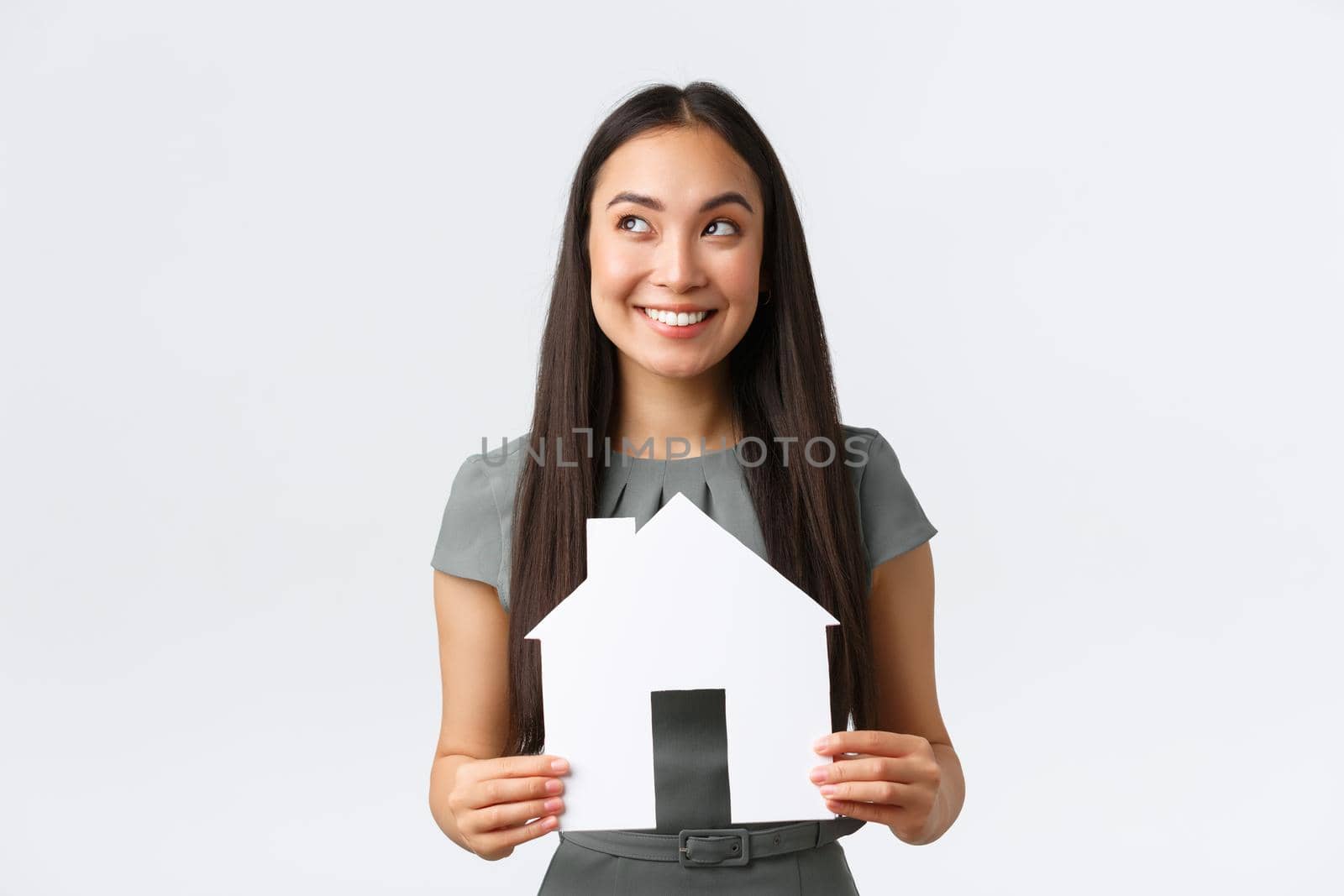 Insurance, loan, real estate and family concept. Thoughtful smiling asian woman in dress, holding paper house in hands and looking away while thinking, imaging perfect home, white background.