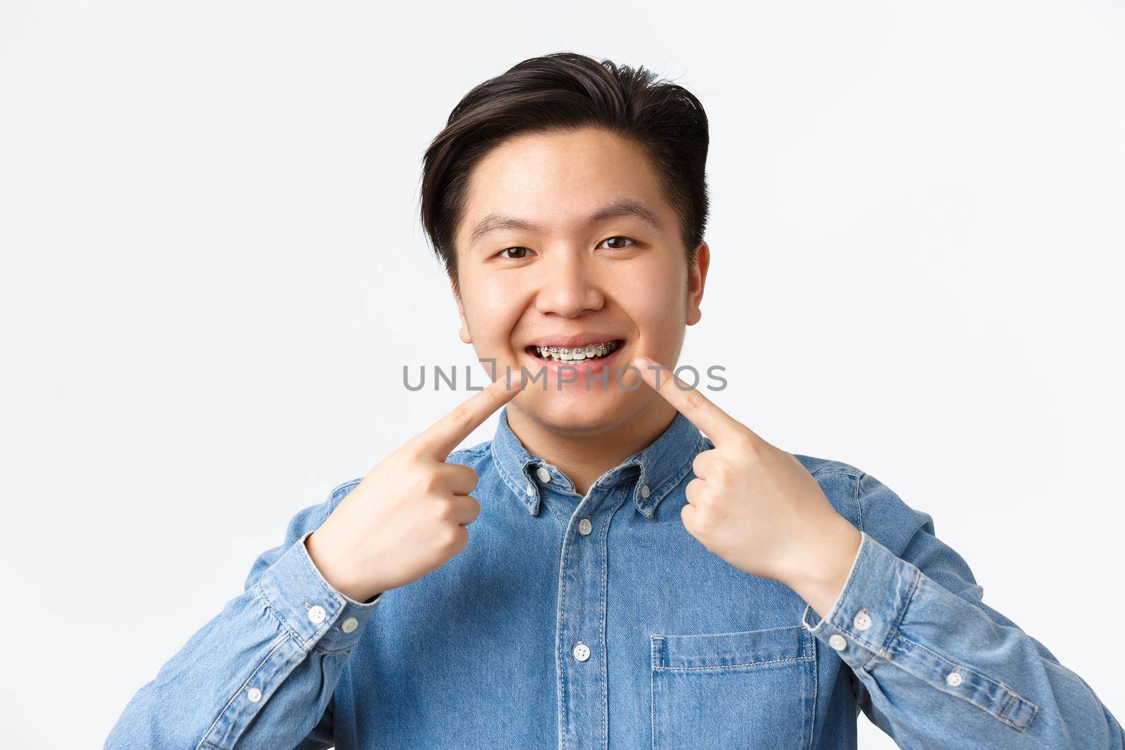 Orthodontics and stomatology concept. Close-up shot of happy smiling asian man recommend dental clinic, pointing fingers at teeth braces with satisfied expression, white background by Benzoix