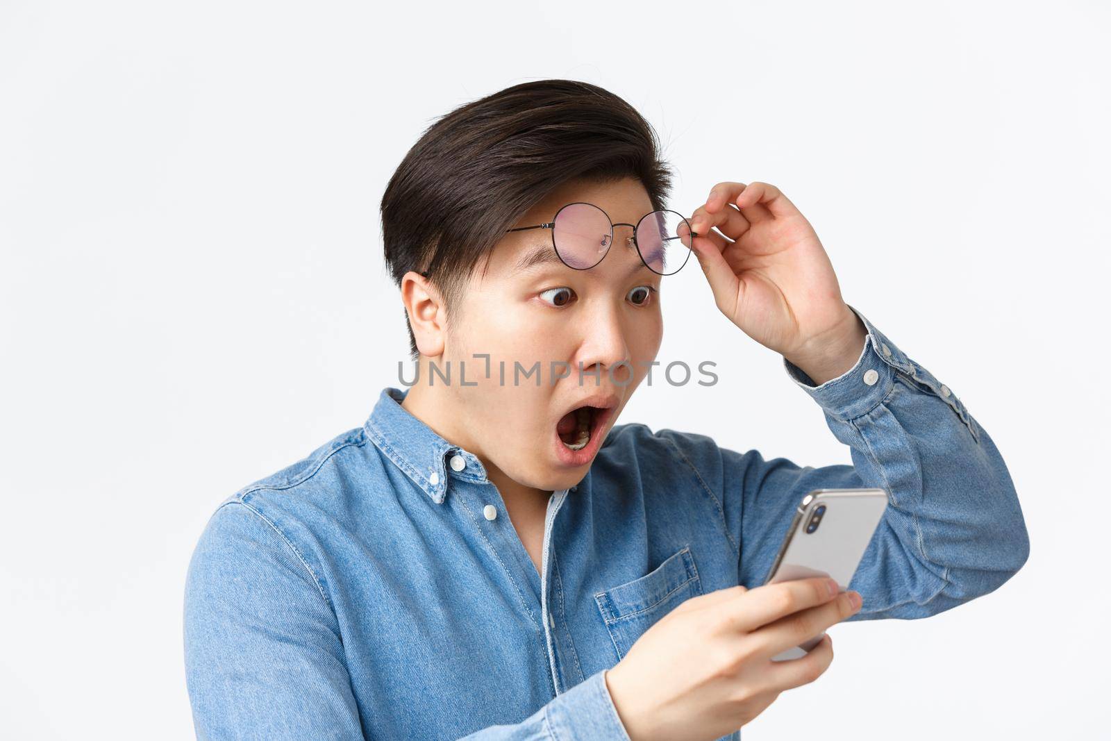 Close-up of shocked asian man take-off glasses, gasping in awe while looking at mobile phone screen, reading shocking news, react to announcement on smartphone, white background.