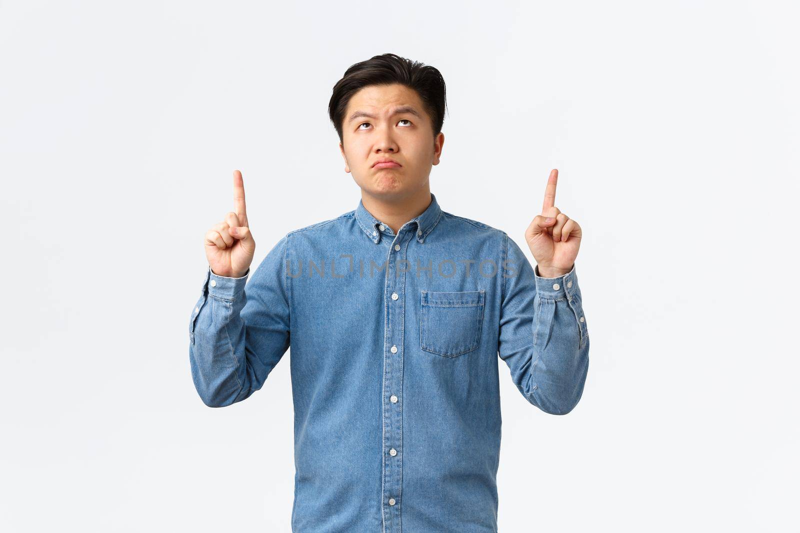 Skeptical and unamused asian man in blue shirt looking and pointing fingers up with disappointed expression, pouting distressed, complaining over something bad, standing white background.