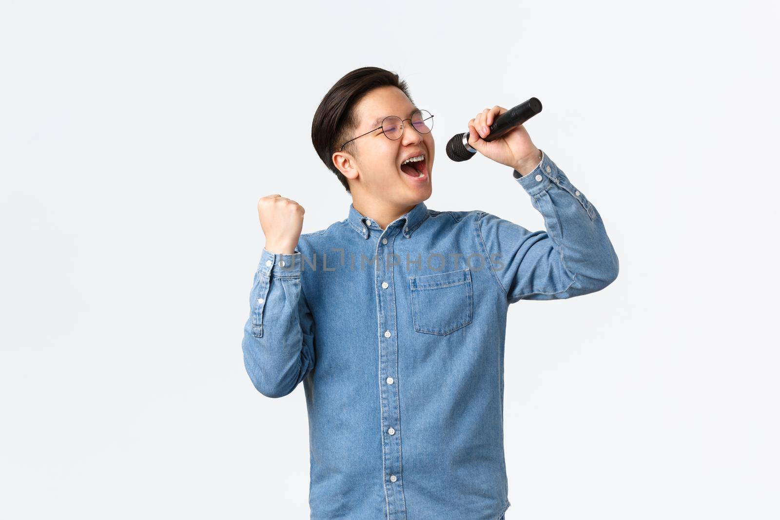 Lifestyle, leisure and people concept. Carefree happy asian man enjoying singing at karaoke, holding microphone and fist pump in delight, performing over white background by Benzoix