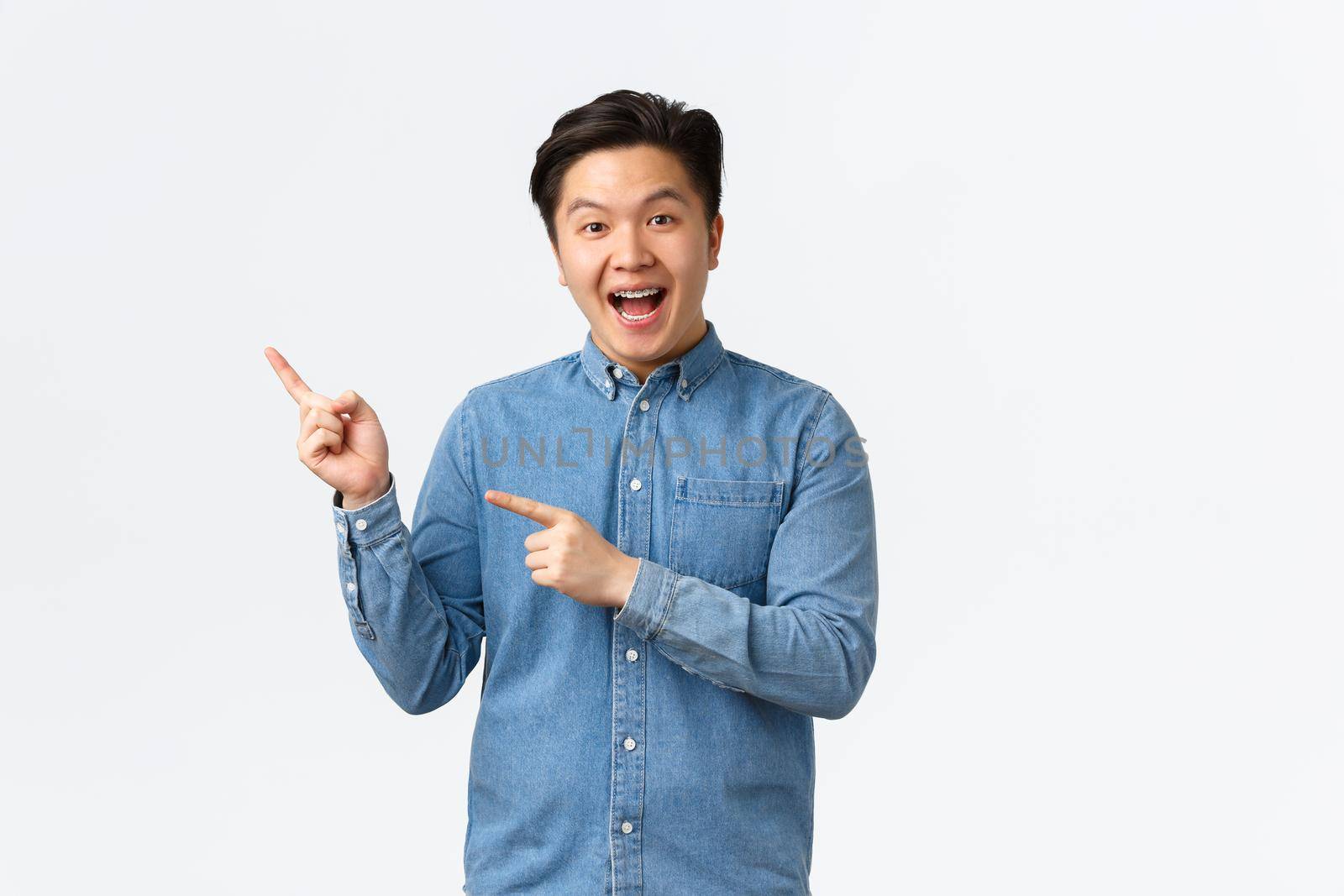 Excited cheerful asian man in blue shirt react to fantastic news, pointing fingers upper left corner, demonstrate banner, promote big event, smiling amazed, standing white background.