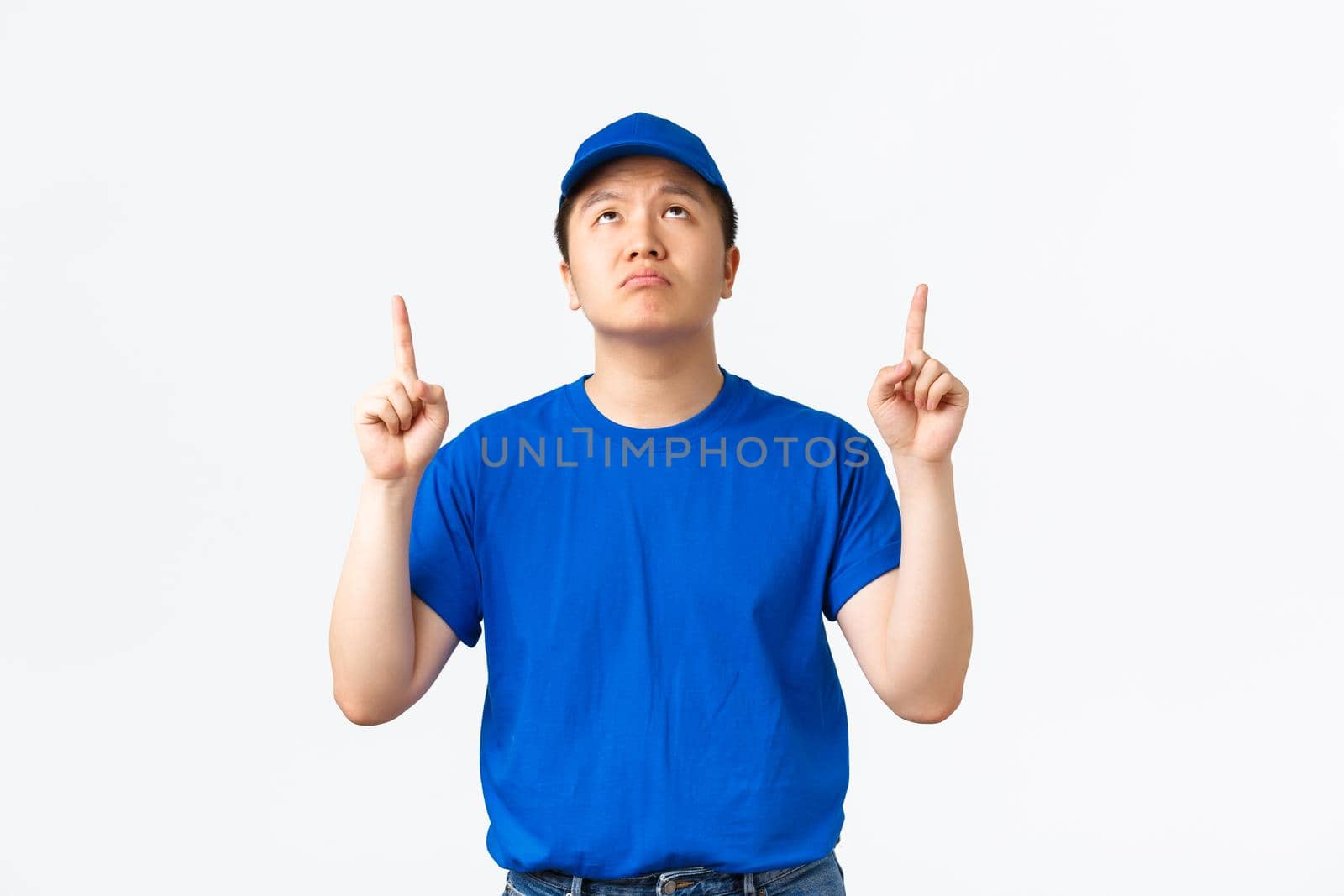 Disappointed and sad asian courier in blue company uniform express regret, frowning and grimacing upset while looking and pointing fingers up, standing white background.