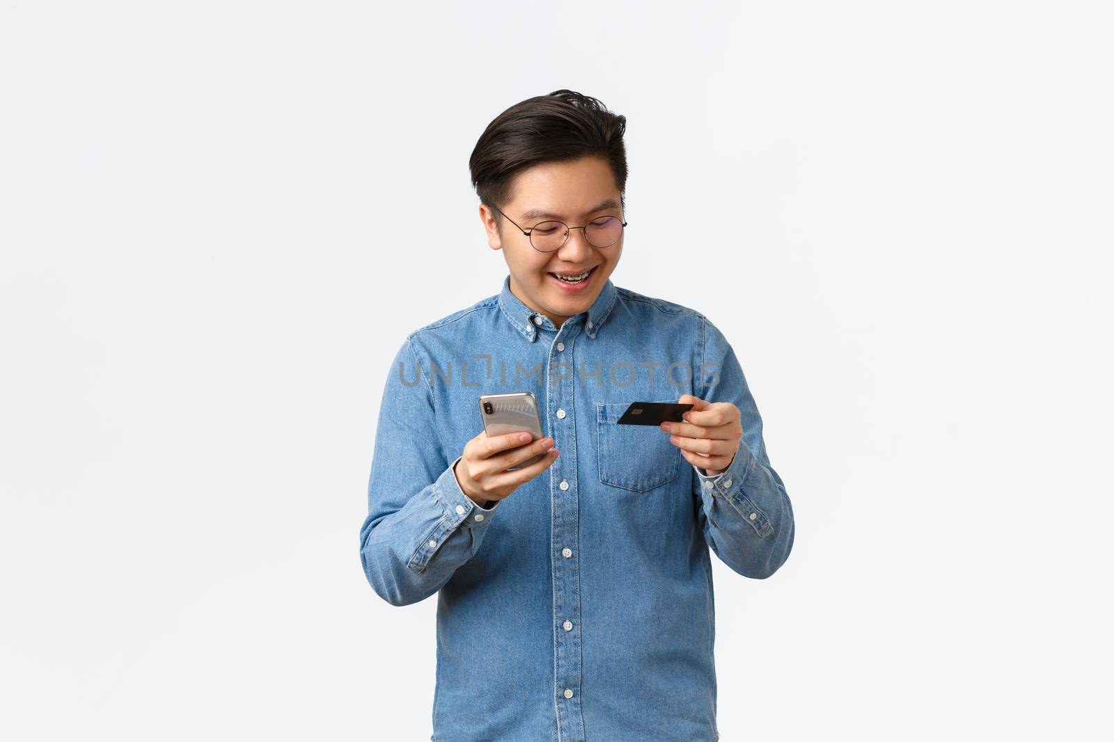 Smiling carefree asian man in glasses and casual clothes, having braces, looking at credit card pleased, buying online with smartphone app, shopping in internet with mobile phone.