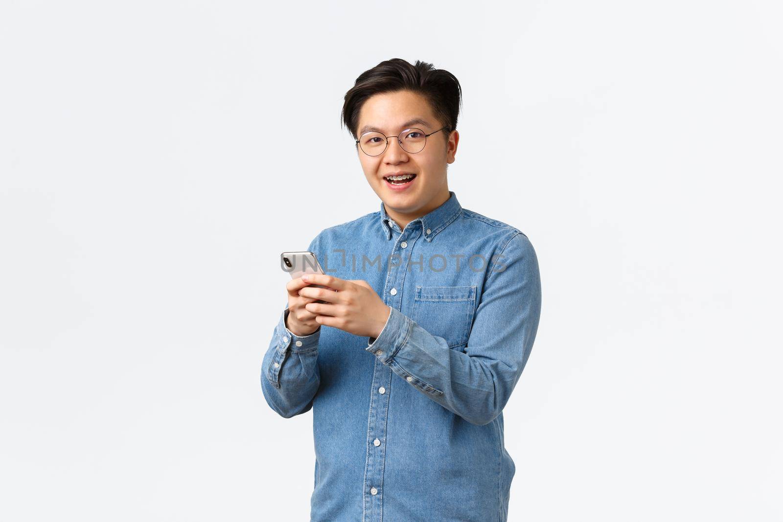 Smiling entertained asian man with braces, wearing glasses, messaging or watching video in internet, using mobile phone application, looking at camera pleased, standing white background.