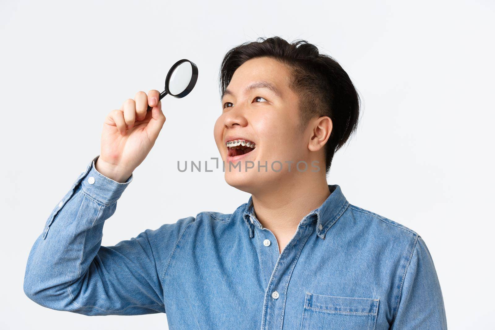 Close-up of amused and excited asian young man with braces, smiling satisfied, found something interesting, looking through magnifying glass at upper left corner, white background by Benzoix