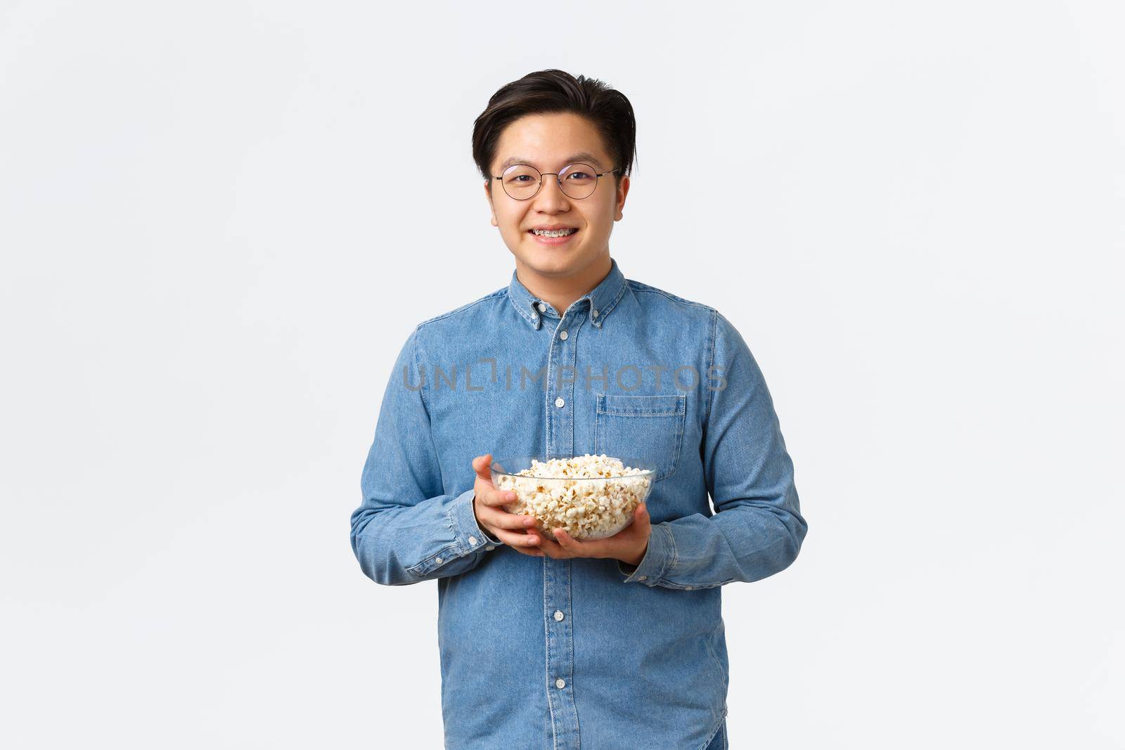 Leisure, lifestyle and people concept. Modest cute asian guy with braces and glasses holding popcorn and smiling at camera, ready for watching premier on tv, standing white background by Benzoix