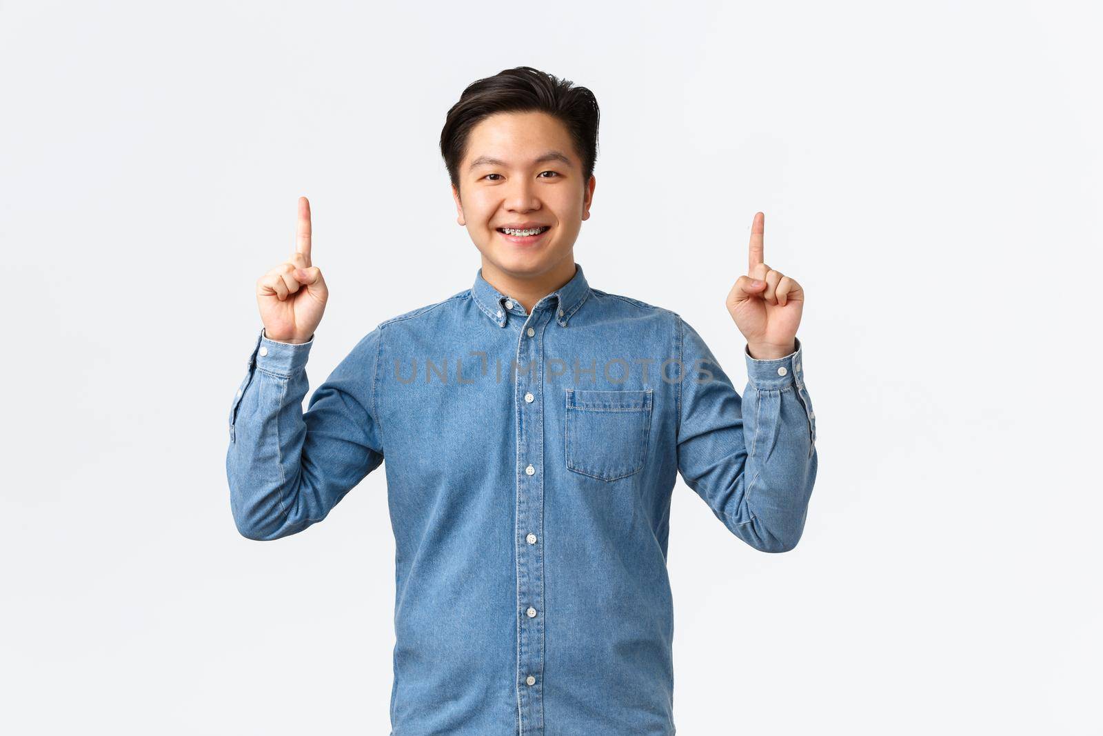 Smiling proud and happy asian male model in blue shirt making announcement, pointing fingers up at banner, recommend buy product, click link, e-commerce and advertisement concept.