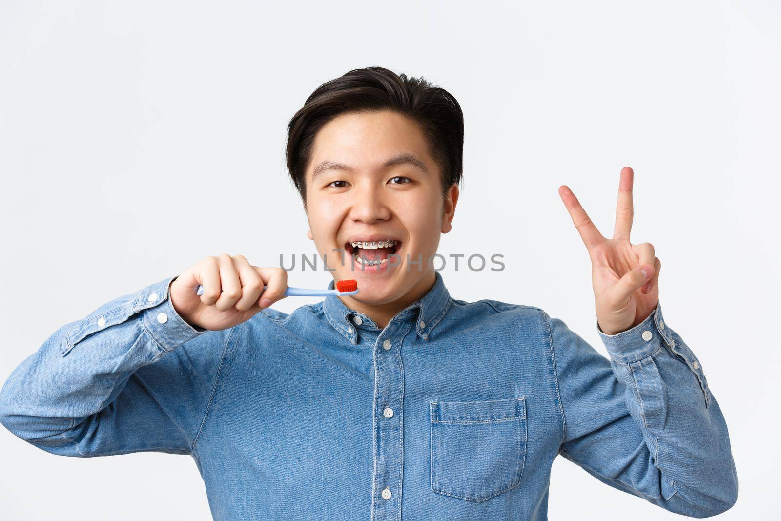 Orthodontics, dental care and hygiene concept. Close-up of happy cute asian man with braces, brushing teeth and smiling, showing peace sign, holding toothbrush, standing white background.