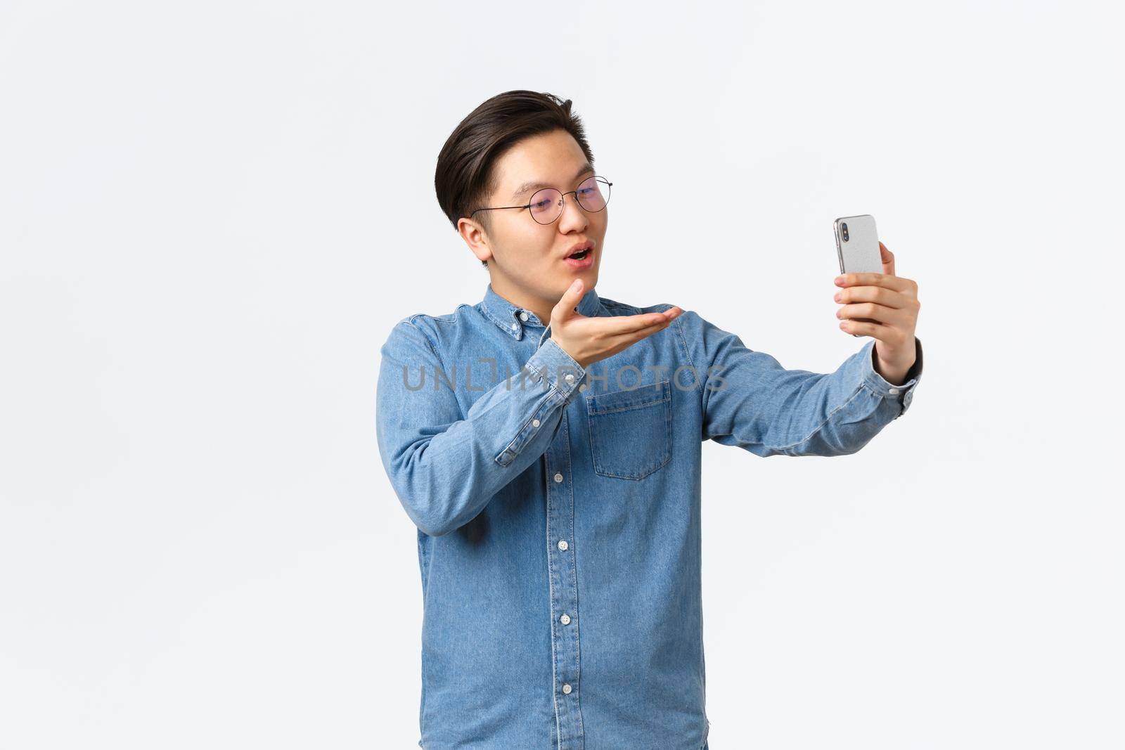 Silly and cute asian guy smiling, talking with girlfriend using smartphone, video calling or taking selfie, sending air kiss at mobile phone camera, standing white background romantic.