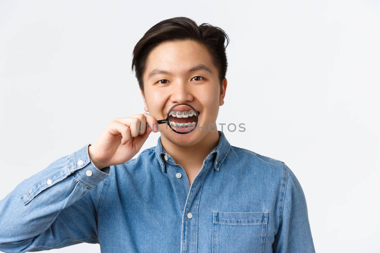 Orthodontics, dental care and stomatology concept. Happy handsome asian young man smiling, open mouth and showing teeth braces with magnifying glass, standing white background by Benzoix