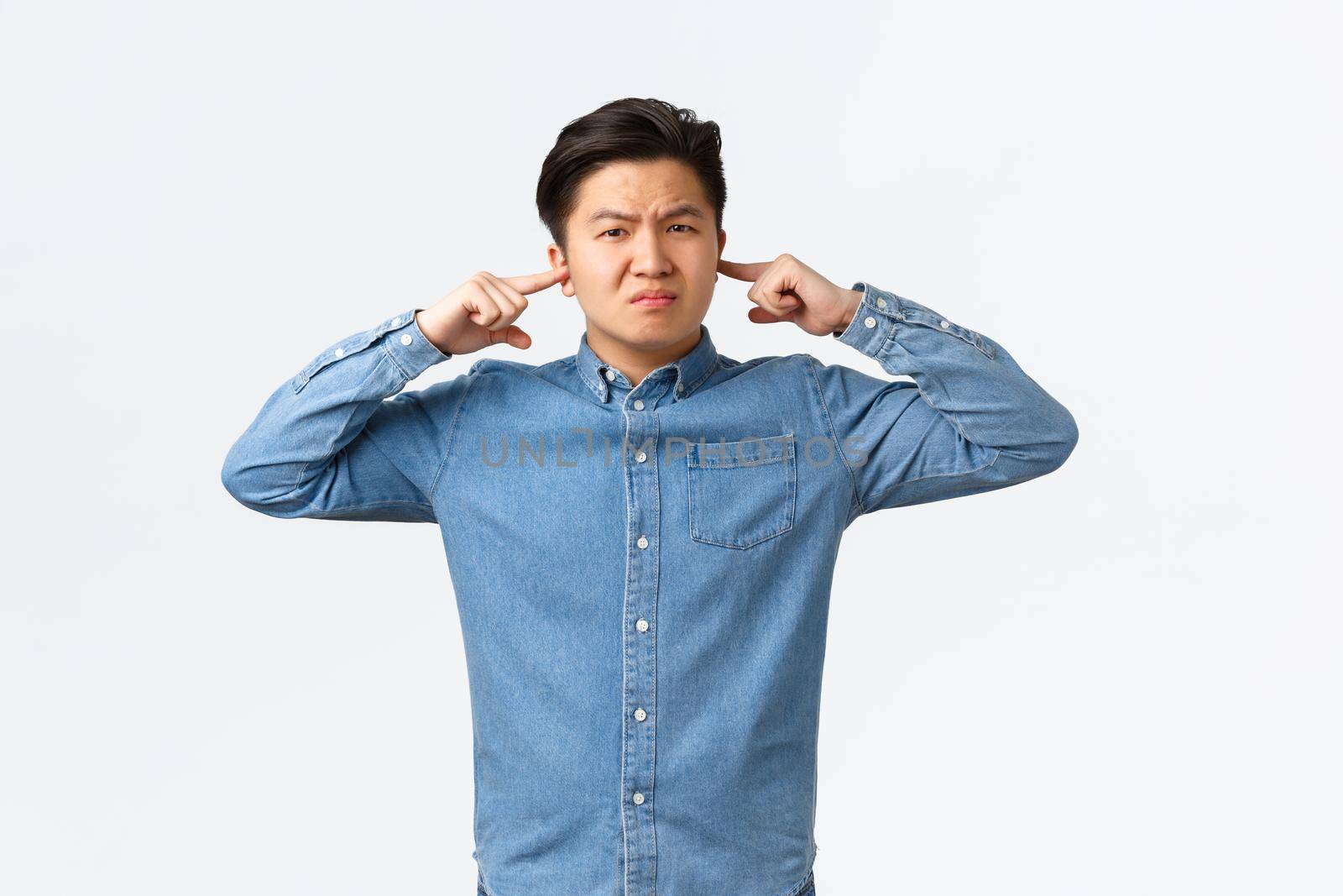 Displeased and bothered asian male student complaining loud neighbour, grimacing displeased, shut ears with fingers and looking annoyed, disturbed with loud sound, standing white background.