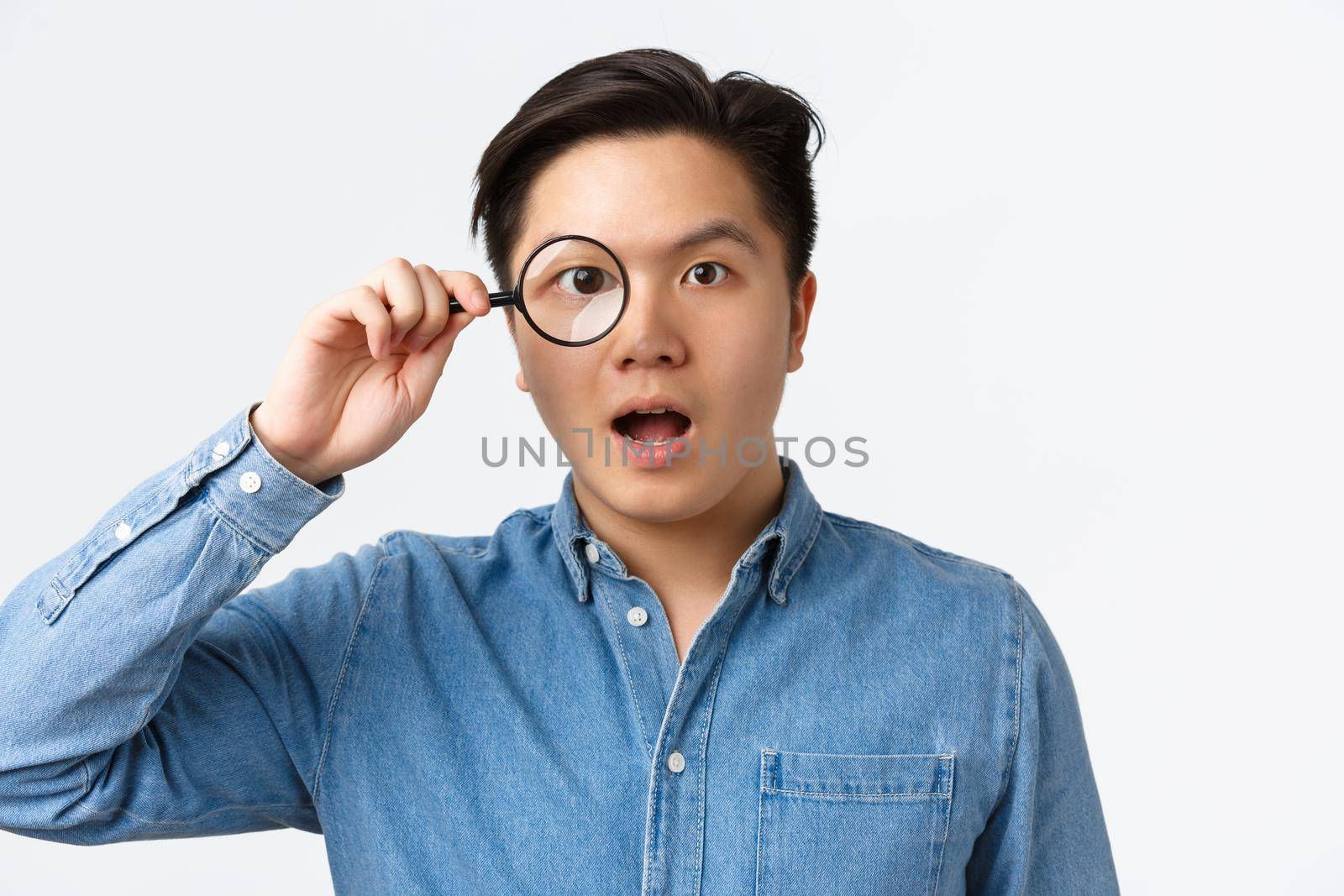 Close-up of funny asian guy looking through magnifying glass and look amazed, searching for something, found interesting product or advertisement, standing white background.