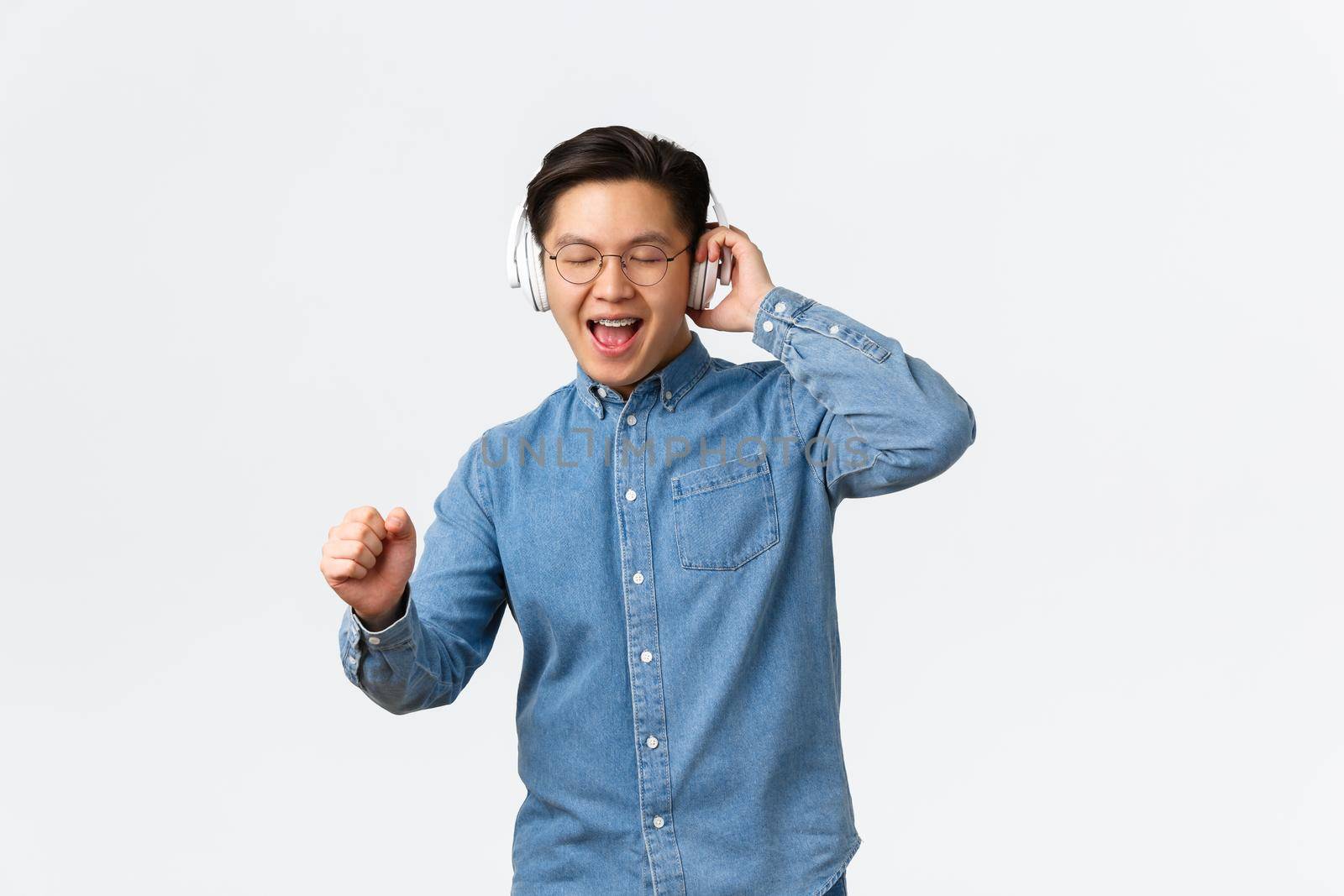 Lifestyle, leisure and technology concept. Happy dancing asian man in glasses, listening music wireless headphones, singing along, having fun with new album on streaming platform by Benzoix