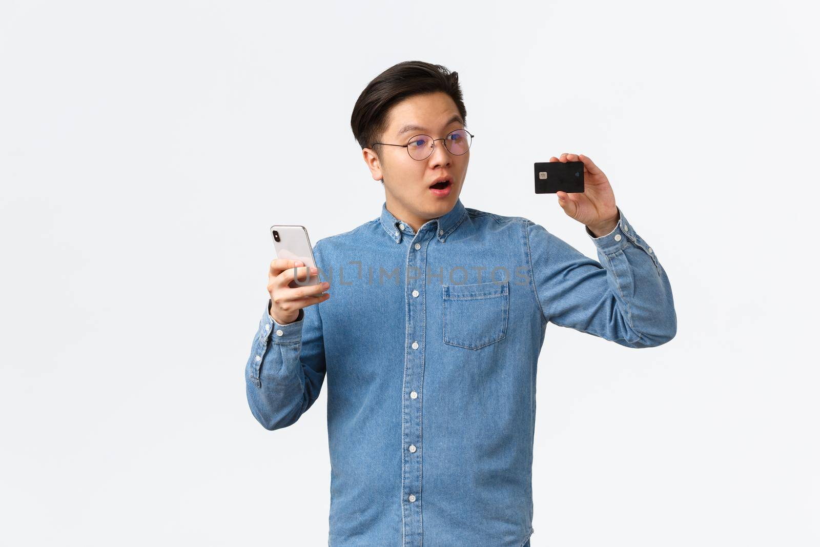 Impressed and fascinated asian guy looking at credit card with amazement, using smartphone, paying for online order, astonished with new e-banking app, standing white background.