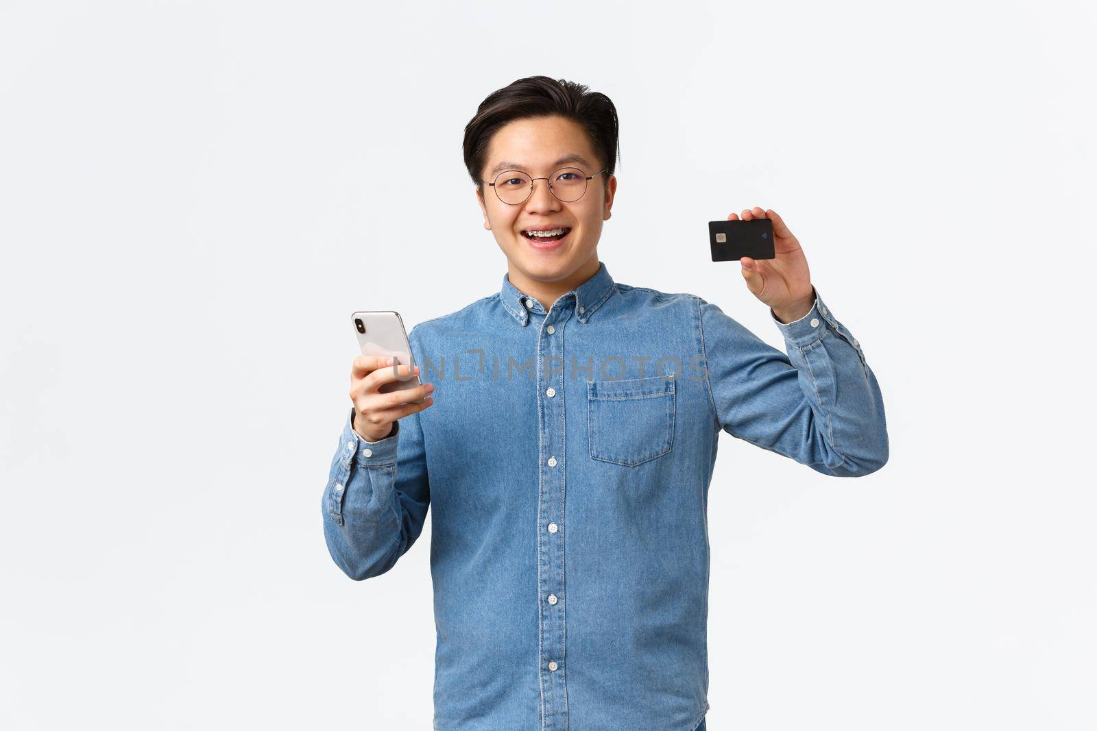 Smiling happy asian guy in glasses and casual clothes using mobile phone to transer money to bank account, showing credit card with satisfied expression, standing pleased white background by Benzoix