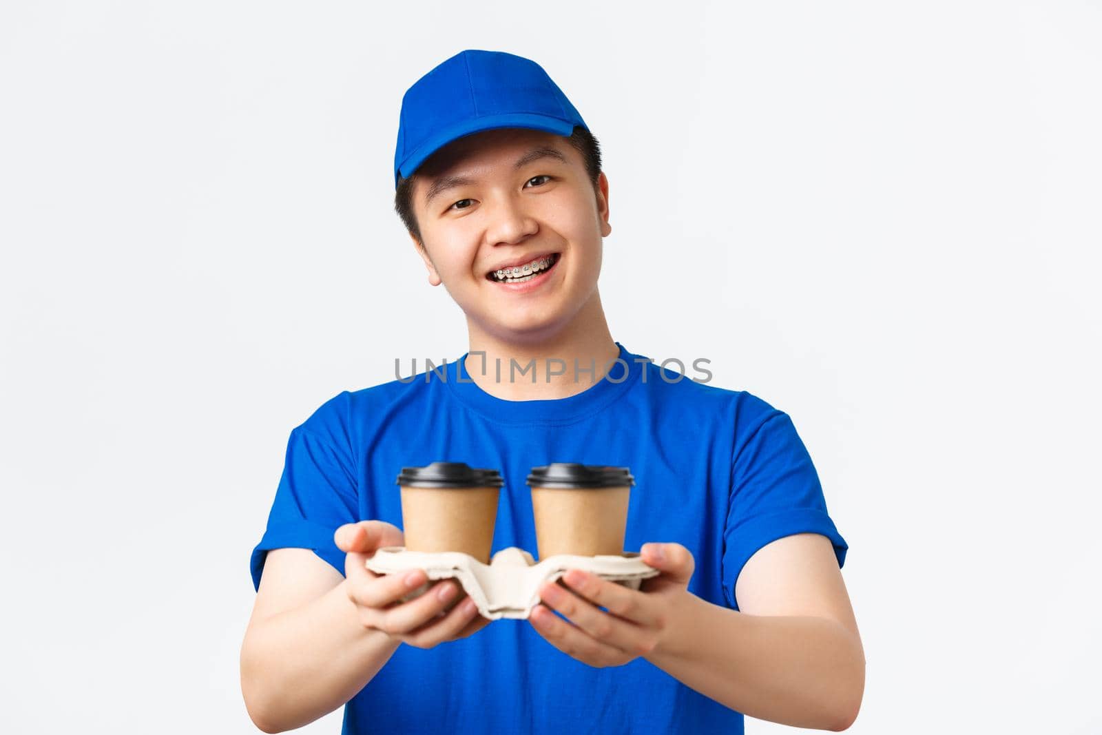 Online orders, takeaway and employees concept. Close-up of cute smiling male courier in blue uniform cap and t-shirt, handing coffee to customer, standing white background.