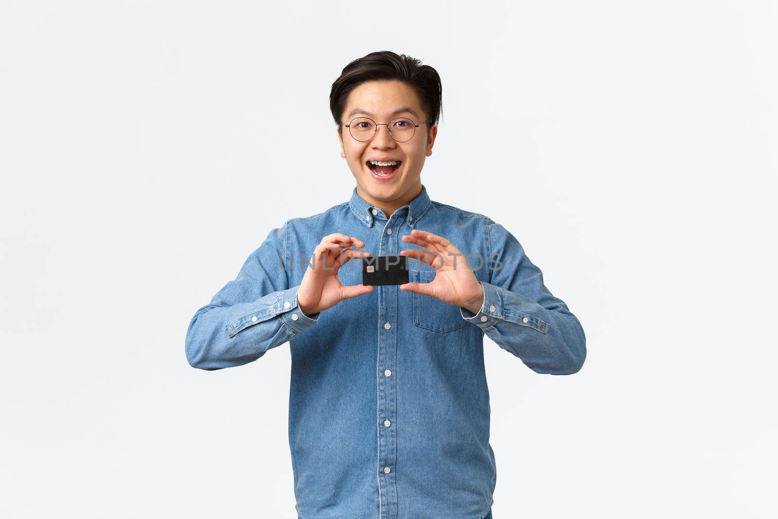 Excited smiling asian guy introduce new banking feature, recommend service, standing in glasses and braces, showing credit card, ready for shopping, paying contactless, white background by Benzoix